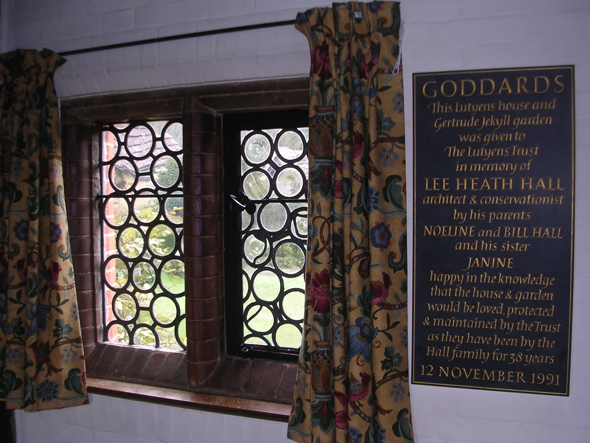 Vista interior de Goddards que muestra una ventana de plomo con paneles circulares, cortinas florales y una placa conmemorativa en la pared