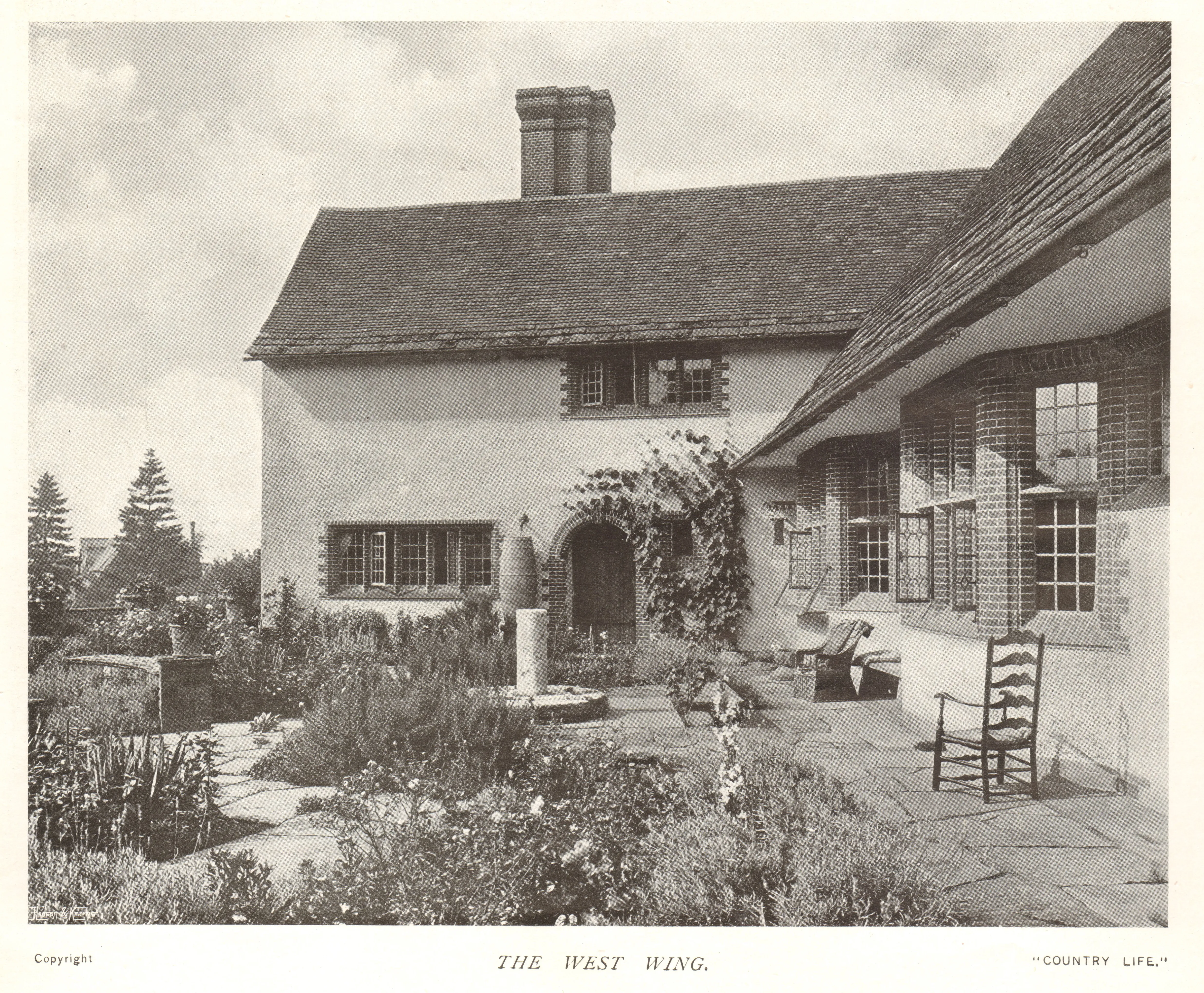 Fotografía histórica en blanco y negro del ala oeste (West Wing) de Goddards que muestra un patio con plantas trepadoras, un pedestal de reloj de sol, sillas de madera y una chimenea.
