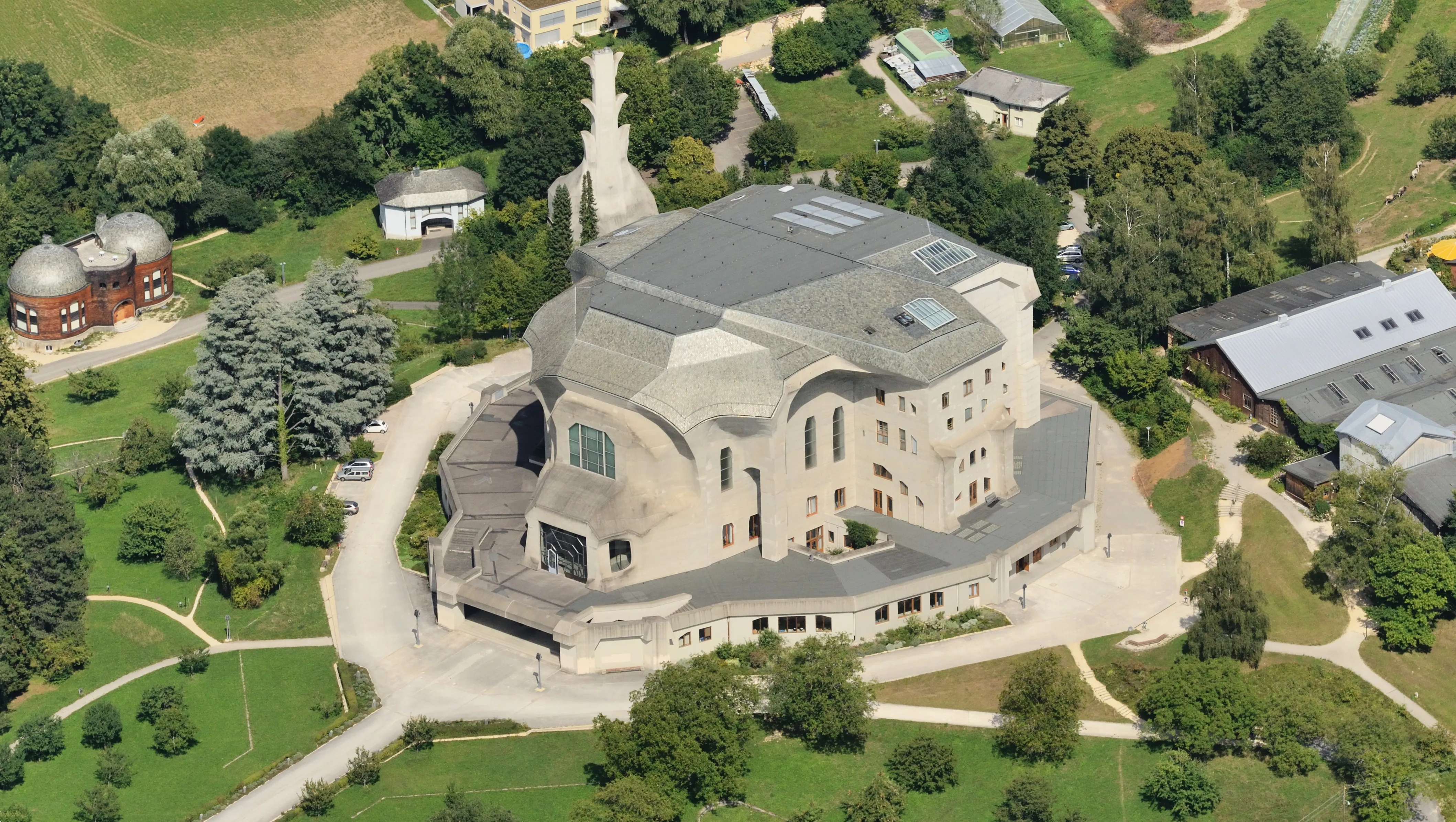 Vista aérea del Goetheanum, un gran edificio de hormigón con un tejado distintivo, rodeado de césped verde, árboles y estructuras más pequeñas.