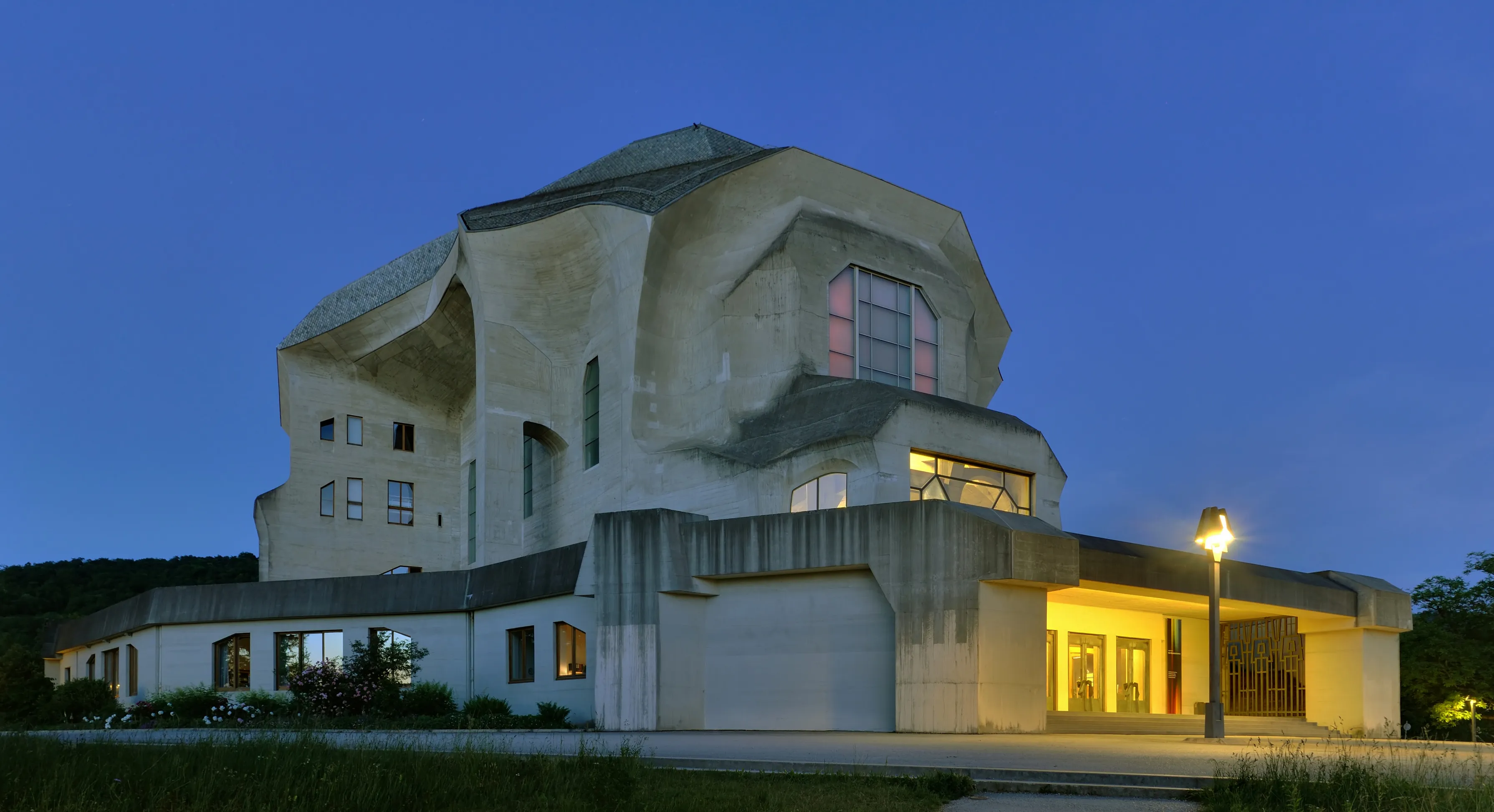 El edificio del Goetheanum en Dornach iluminado al atardecer, mostrando su distintiva fachada de hormigón escultural contra un cielo crepuscular azul intenso