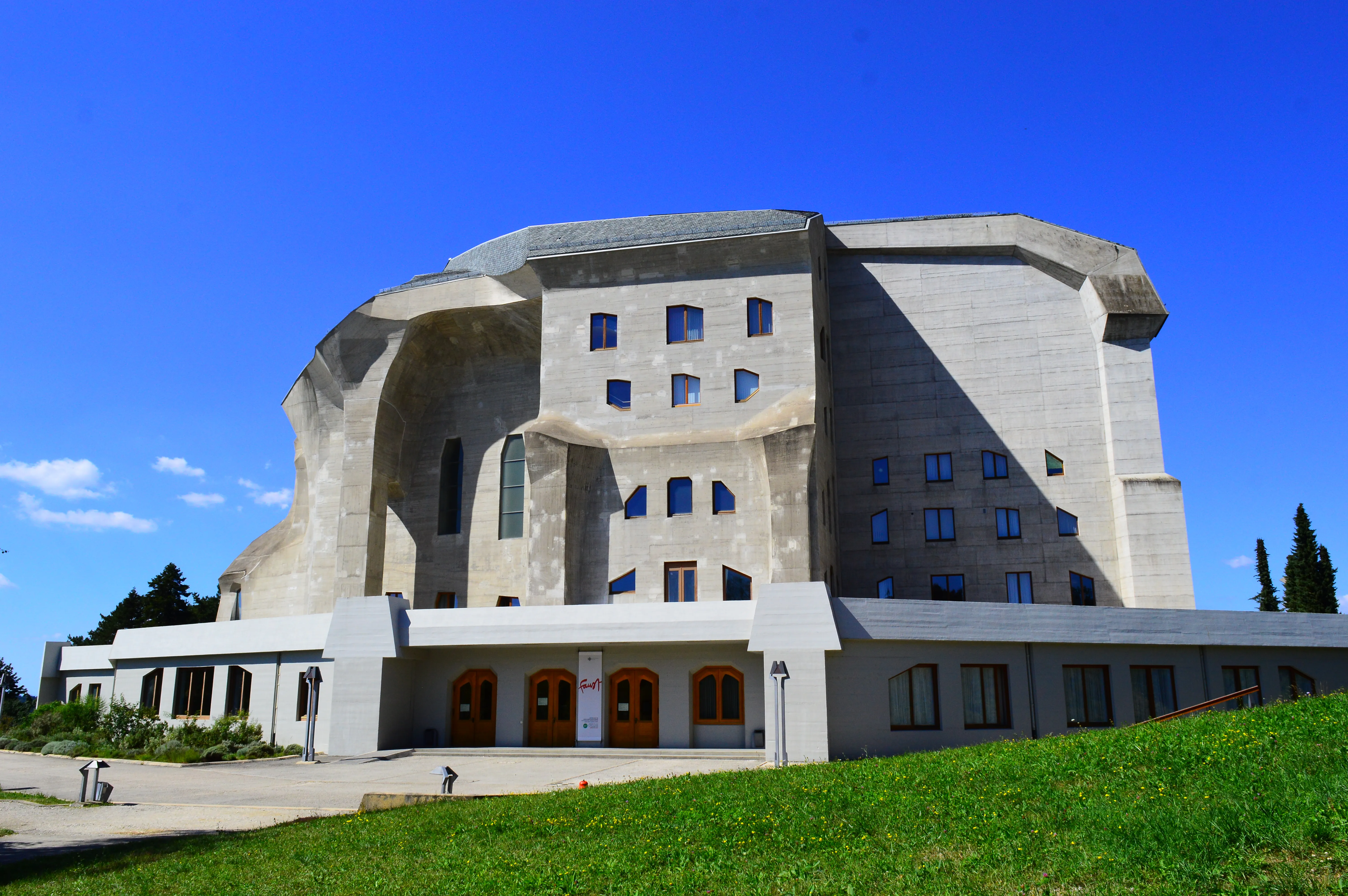 Plano general del Goetheanum, un gran edificio de hormigón con una arquitectura distintiva, curva y angulosa, sobre un cielo azul despejado y con un césped verde en primer plano.