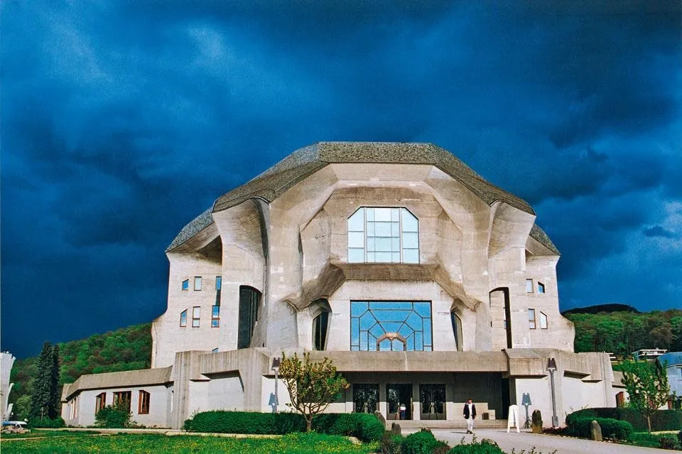 Vista exterior del edificio del Goetheanum en Dornach, que presenta su arquitectura de hormigón única con grandes ventanas y una línea de techo distintiva sobre un espectacular cielo azul