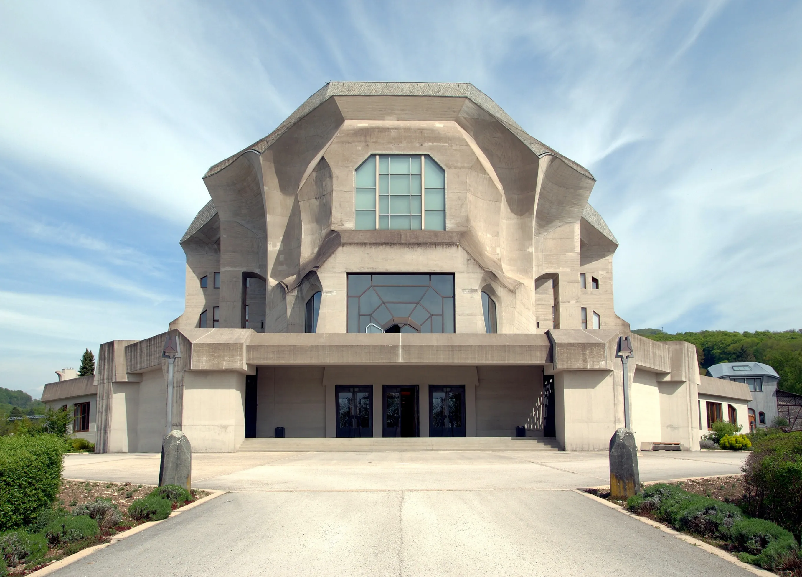 Fachada frontal del Goetheanum en Dornach, un gran edificio de hormigón curvo con una ventana central de múltiples paneles, puertas de entrada bajo un amplio voladizo y un camino pavimentado que conduce hacia él bajo un cielo azul con nubes tenues