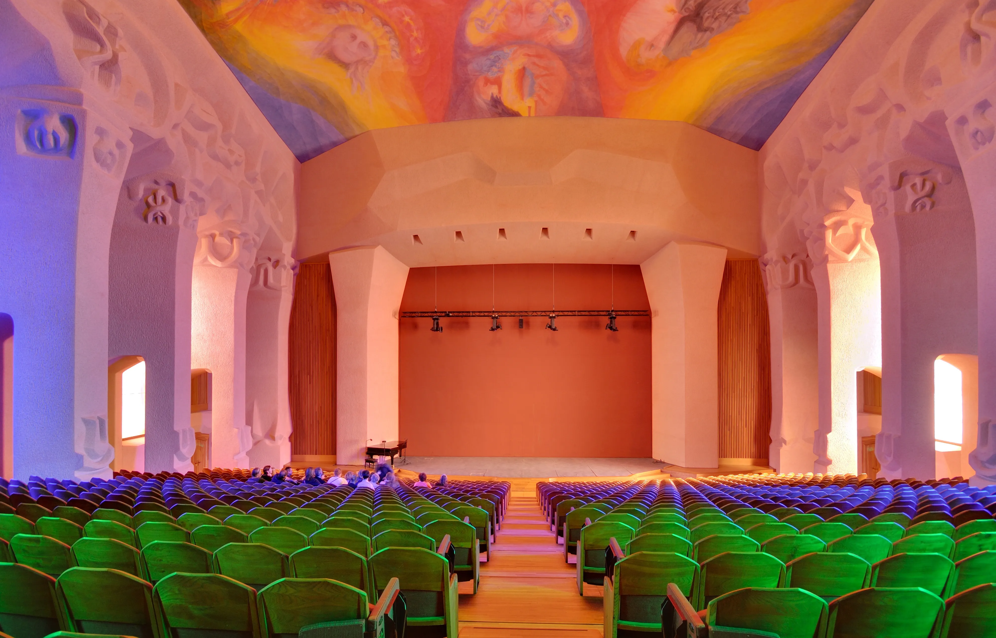Vista interior de un gran auditorio con filas de asientos verdes frente a un escenario, flanqueado por columnas escultóricas blancas y un techo pintado de colores.