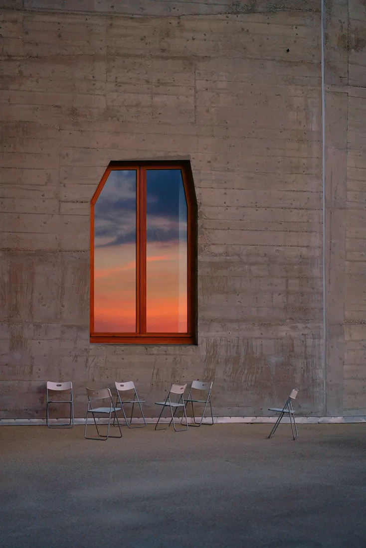 Pared interior de hormigón con una ventana arqueada de marco rojo que muestra el cielo al atardecer y sillas plegables dispuestas debajo