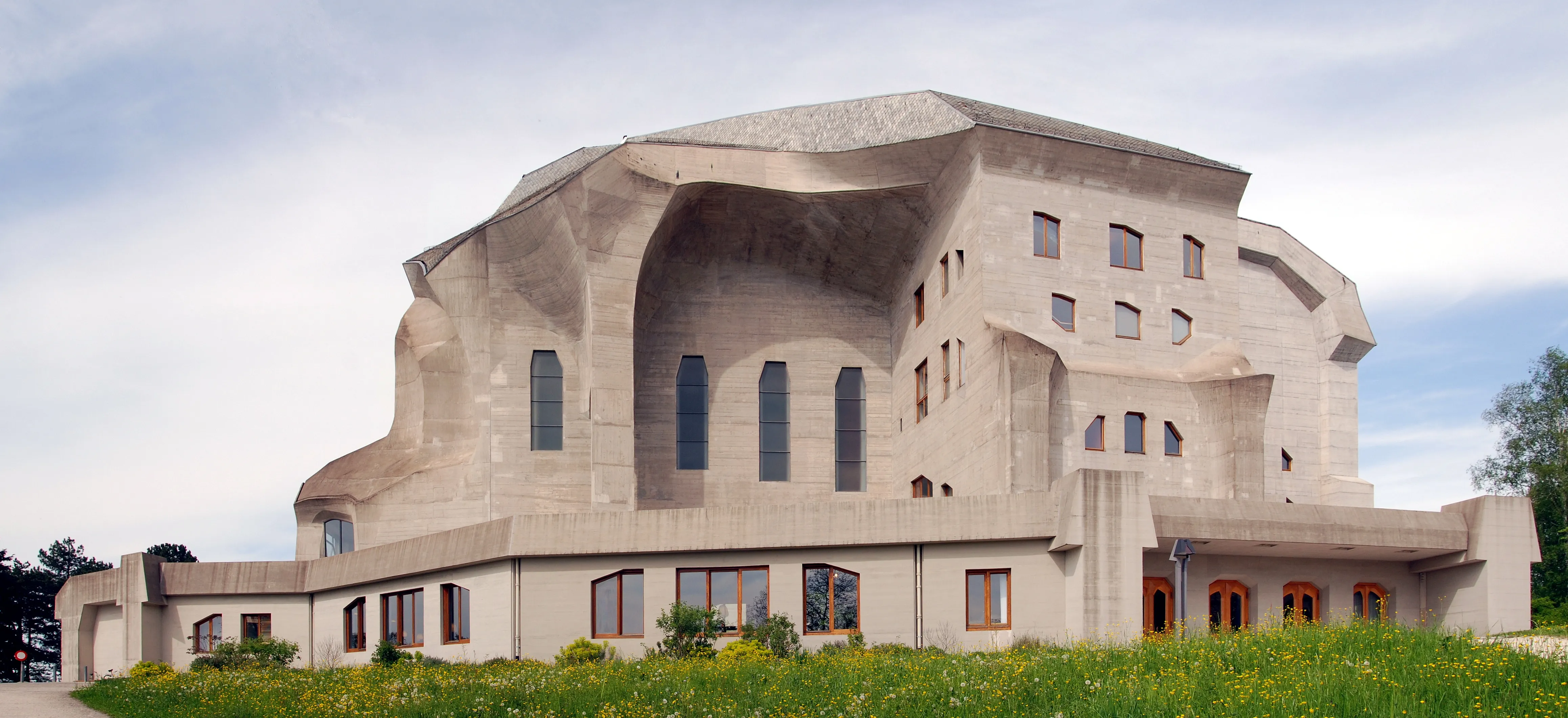 Vista exterior del Goetheanum, un gran edificio de hormigón escultural con formas orgánicas y ventanas altas y estrechas, rodeado de césped verde y árboles bajo un cielo nublado.