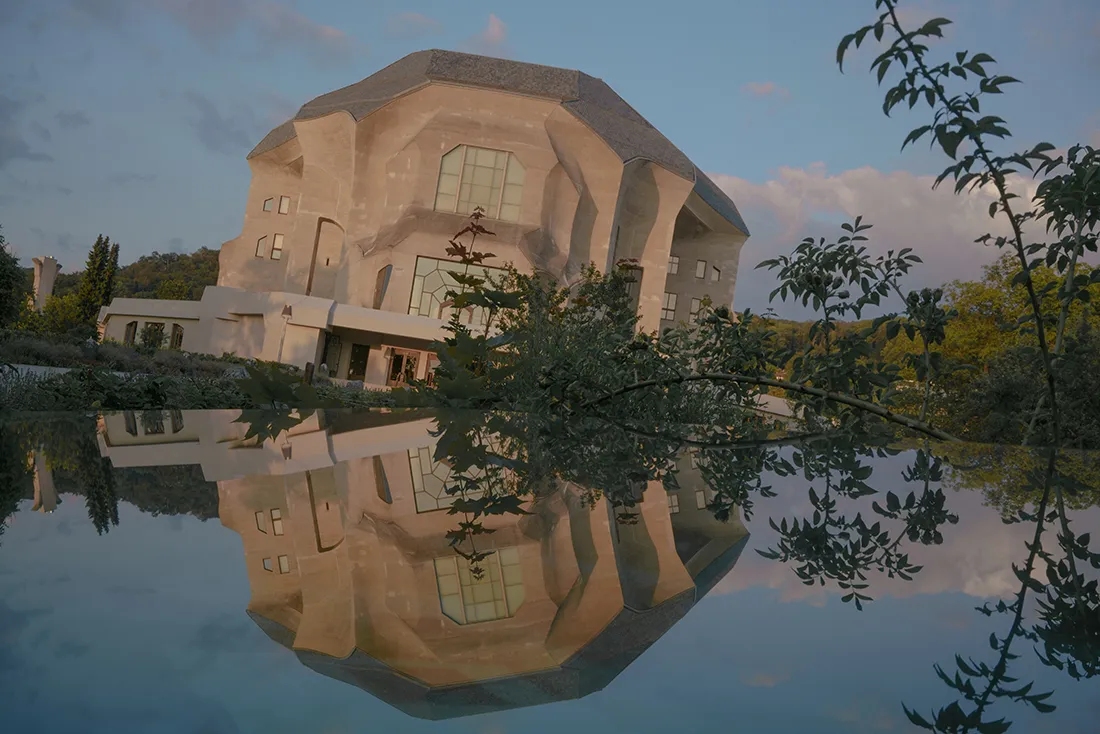 El edificio del Goetheanum reflejado en un estanque tranquilo al atardecer, con ramas de árboles enmarcando el lado derecho y la fachada de hormigón reflejada en el agua.