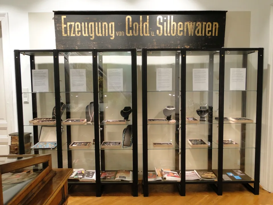 Vitrina de cristal con artefactos y herramientas de metal, con un cartel encima que dice 'Erzeugung von Gold- u. Silberwaren'