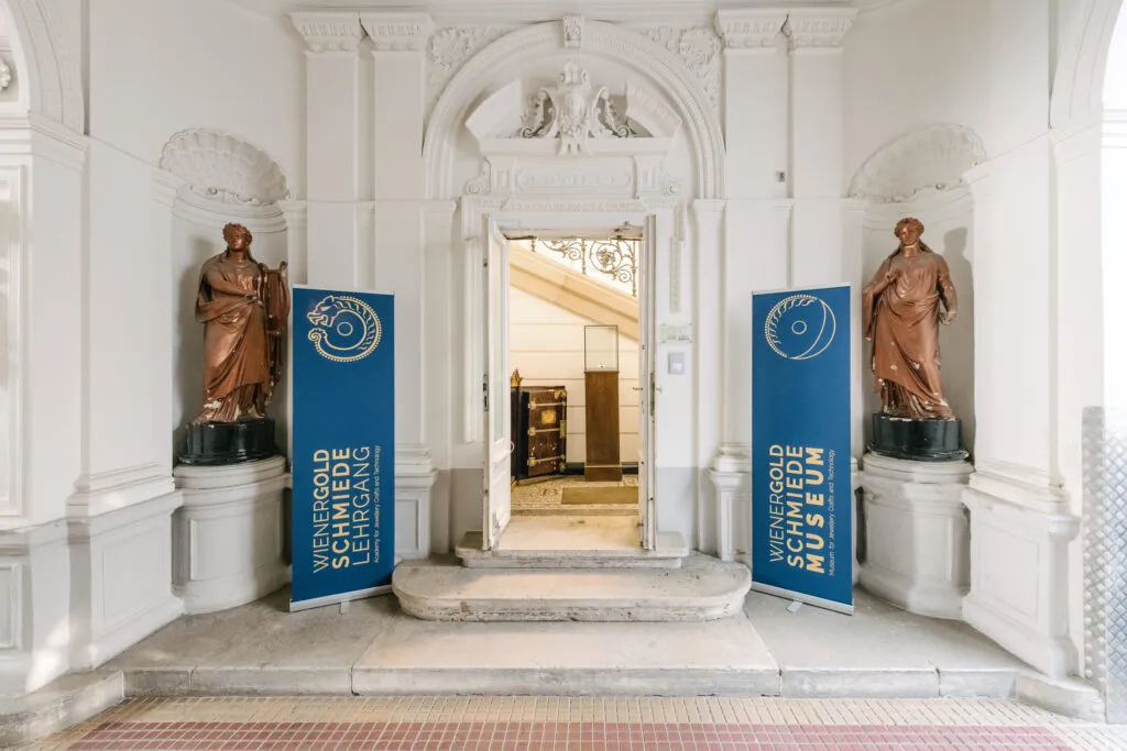 Vestíbulo con dos estatuas de bronce sobre pedestales, pancartas azules con texto y una puerta que conduce a un espacio interior.