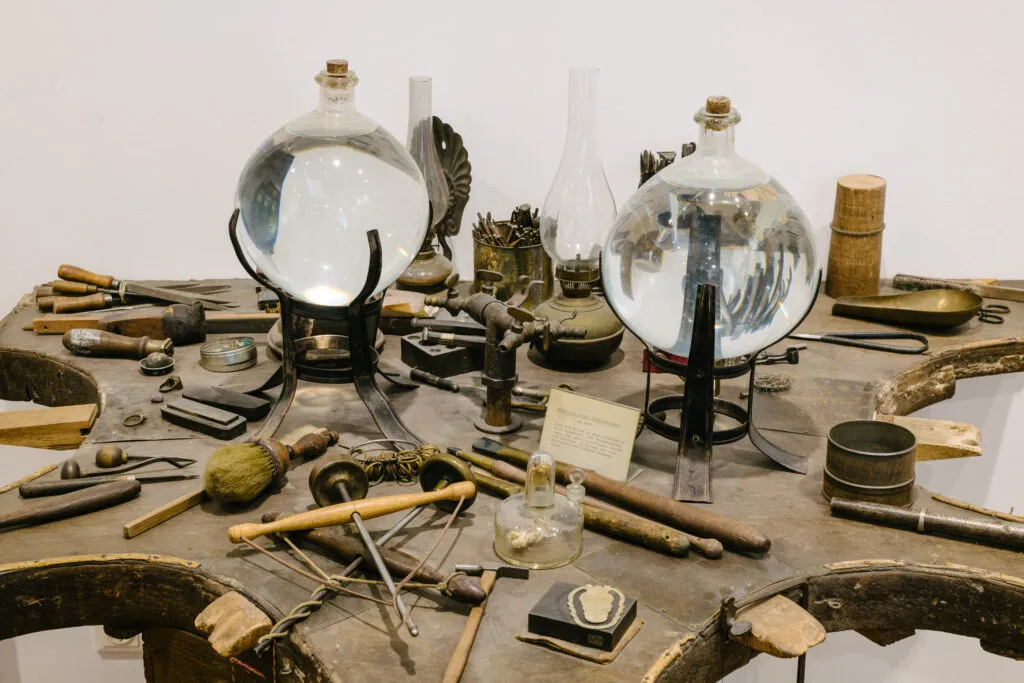Una colección de herramientas históricas de orfebrería y platería sobre un banco de trabajo de madera con dos grandes recipientes de vidrio sobre soportes