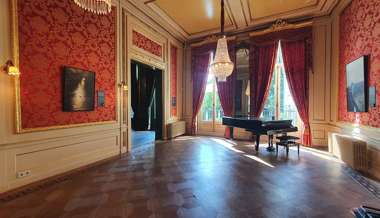 Gran sala con paredes ornamentadas de patrón rojo, candelabros y un piano de cola en el Grachtenmuseum, Ámsterdam