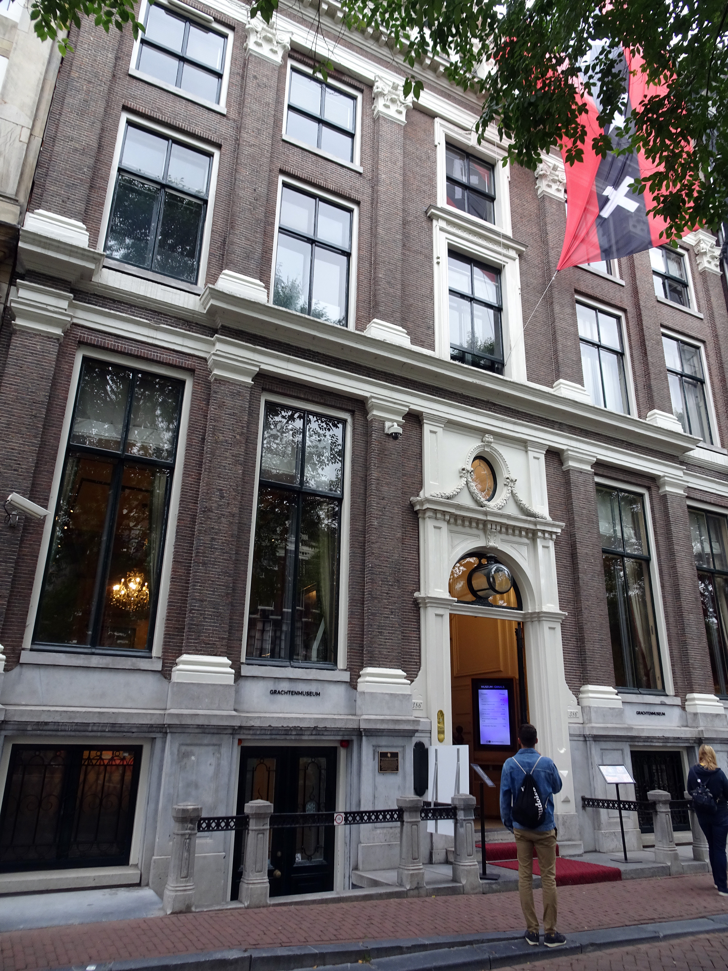 Fachada de casa en un canal de Ámsterdam con paredes de ladrillo, molduras blancas y grandes ventanas. Entrada con pantalla digital, alfombra roja y peatones. Bandera roja y negra con cruz blanca encima.