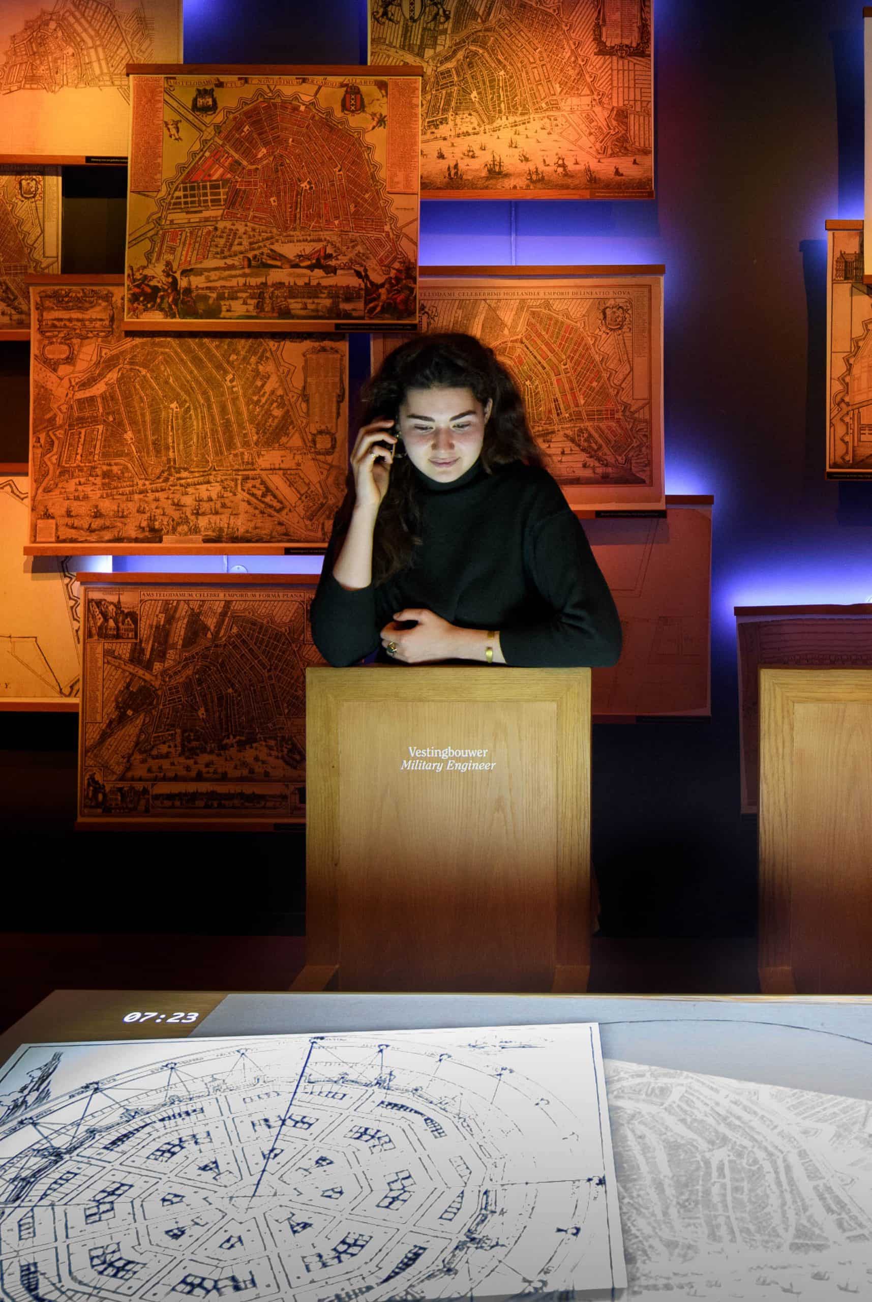 Zwiedzający przy drewnianym podium w słabo oświetlonej galerii muzealnej z historycznymi mapami kanałów na ścianach i planami architektonicznymi na stole