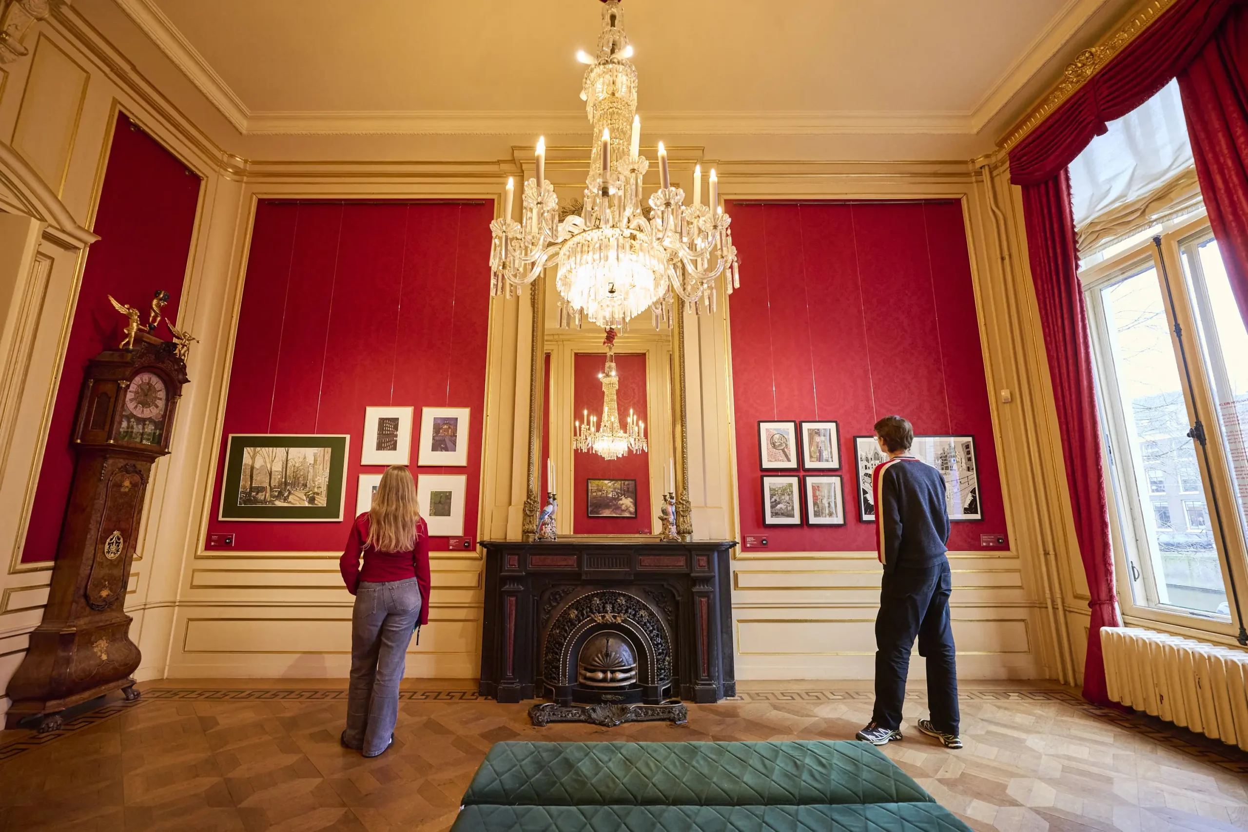 Sala de museo de paredes rojas con candelabro de cristal, fotografías enmarcadas, chimenea negra y dos visitantes examinando exposiciones cerca de altos ventanales con cortinas rojas