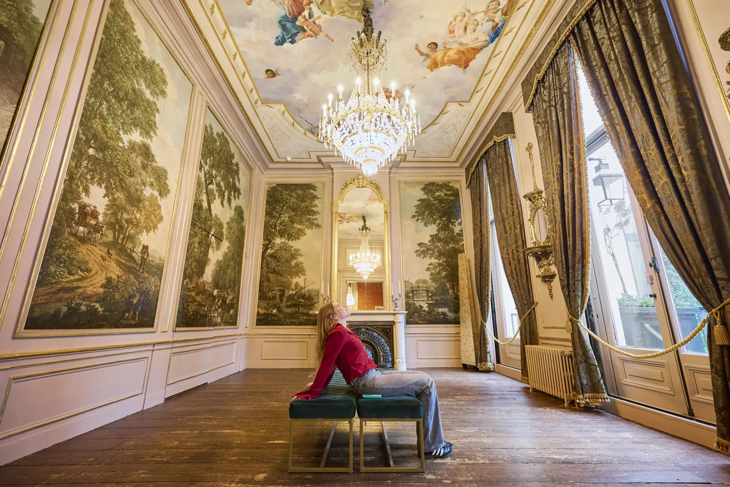 Ornada sala de museo con frescos pintados en el techo, grandes murales de paisajes, candelabros de cristal y una persona sentada en un banco cerca de altos ventanales.