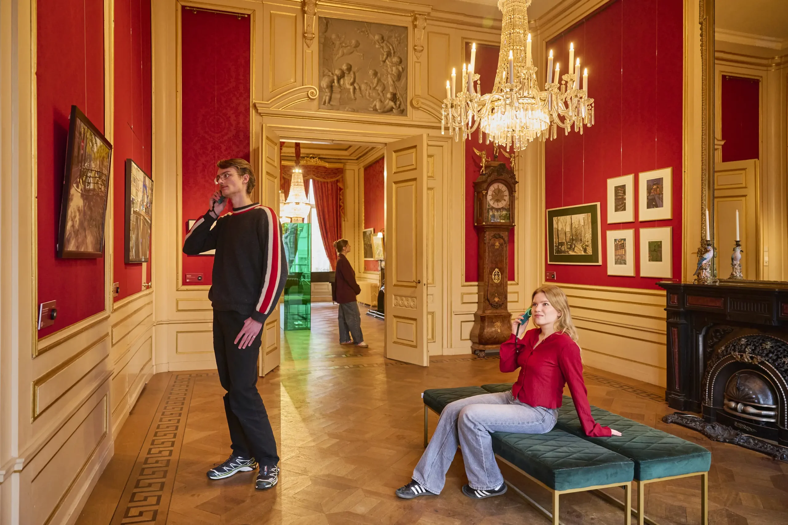 Gran sala con paredes rojas, adornos dorados y candelabro. Dos personas en el espacio, una de pie, otra sentada en un banco verde. Se aprecian pinturas y un reloj de pie.