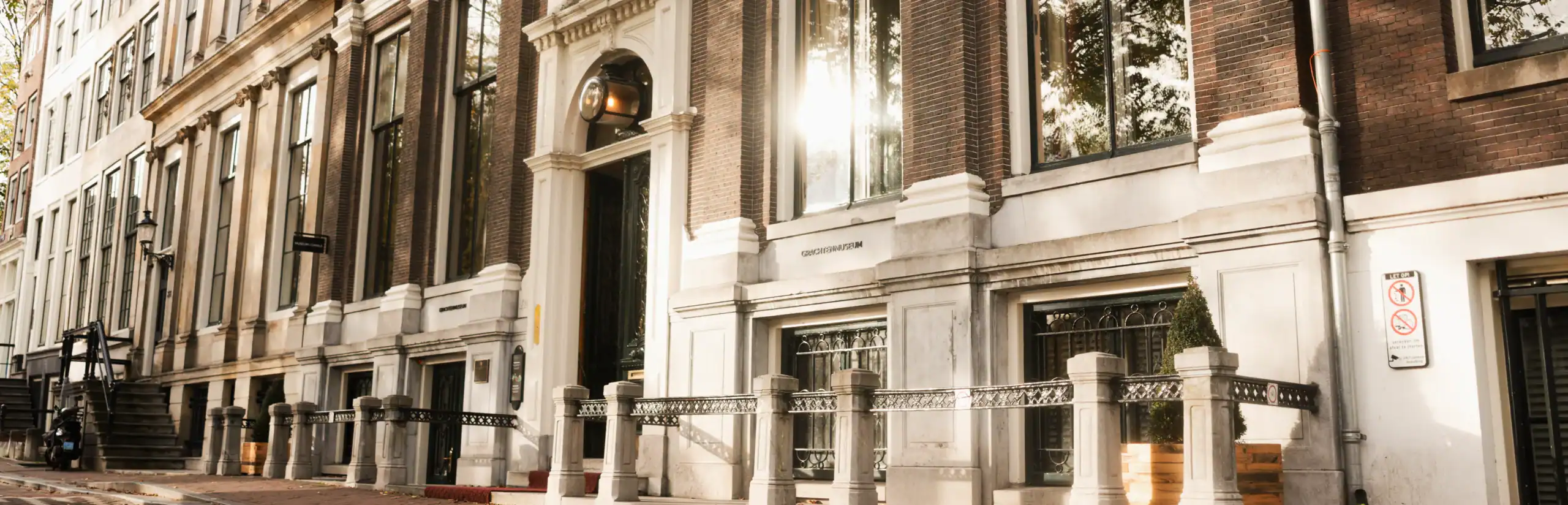 Fachada histórica de casa en un canal con ventanas altas, entrada de piedra y detalles arquitectónicos ornamentados bajo la luz del sol, con bolardos en la calle y edificios adyacentes