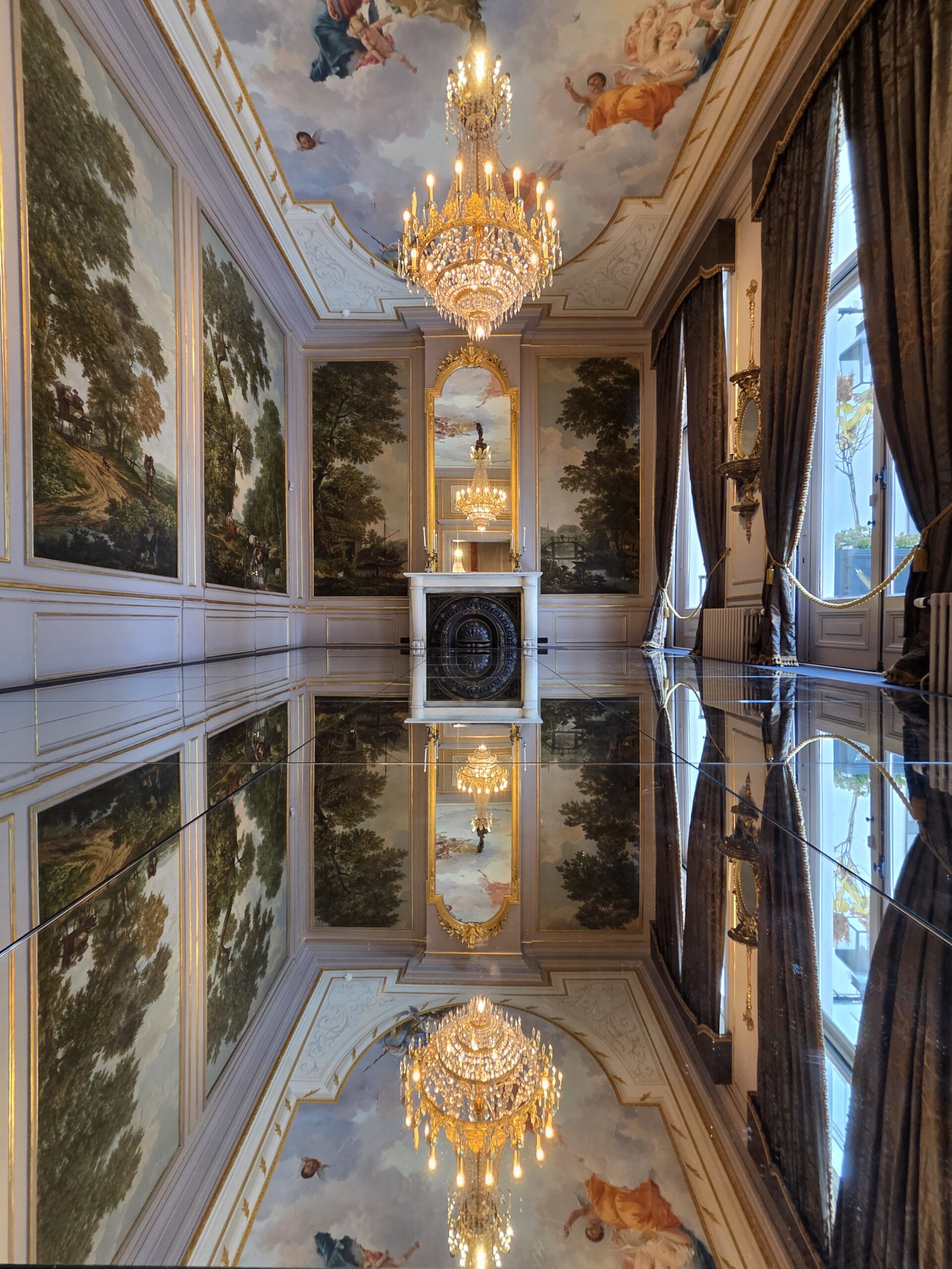Opulenta sala de museo con candelabros dorados, paneles de pared pintados con paisajes, suelo de mármol reflectante y frescos en el techo que representan figuras.