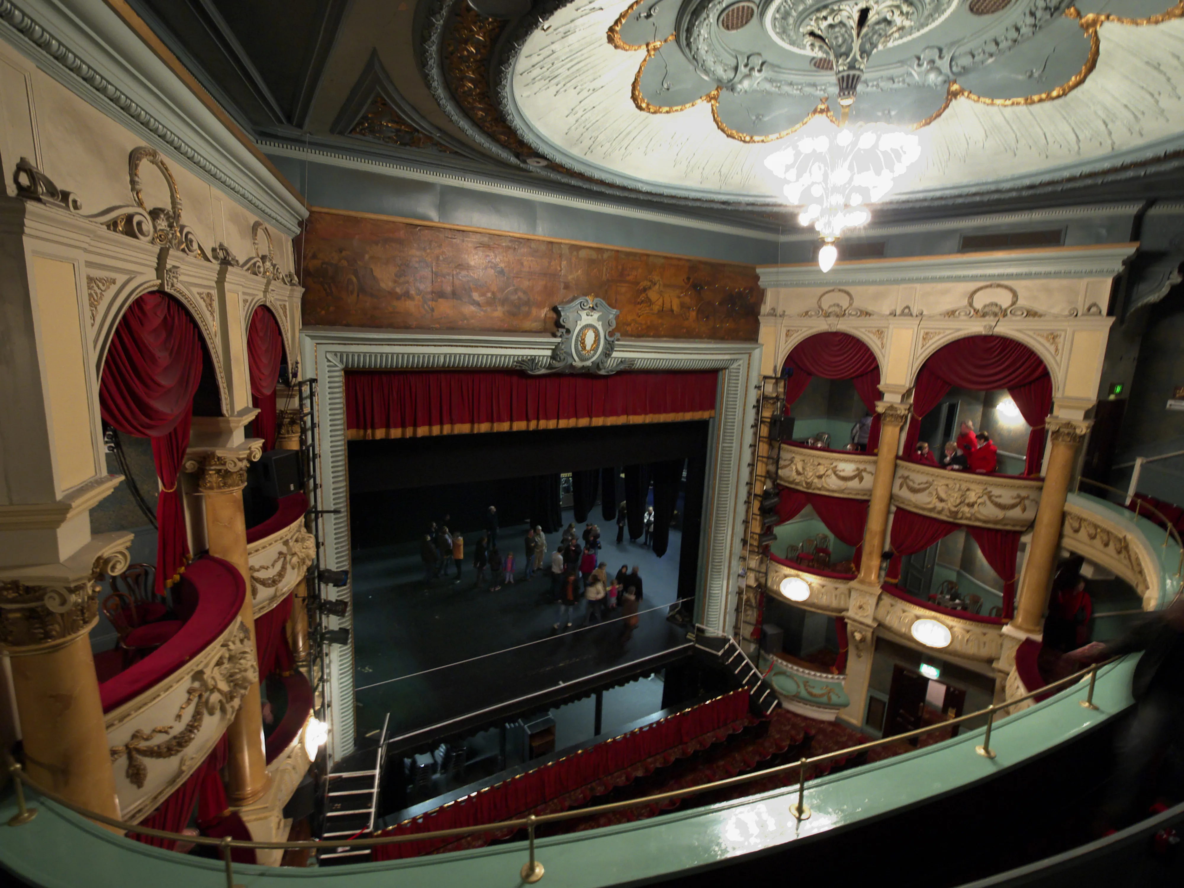 Rijk versierd theaterinterieur met balkons van rood fluweel, decoratieve zuilen van pleisterwerk, een kristallen kroonluchter en een podium met mensen beneden.