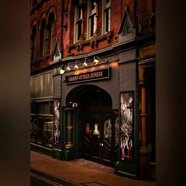 Nachtelijk aanzicht van de ingang van het Grand Opera House met een verlicht bord boven de boogvormige deur, een roodbakstenen gevel en theateraffiches aan weerszijden