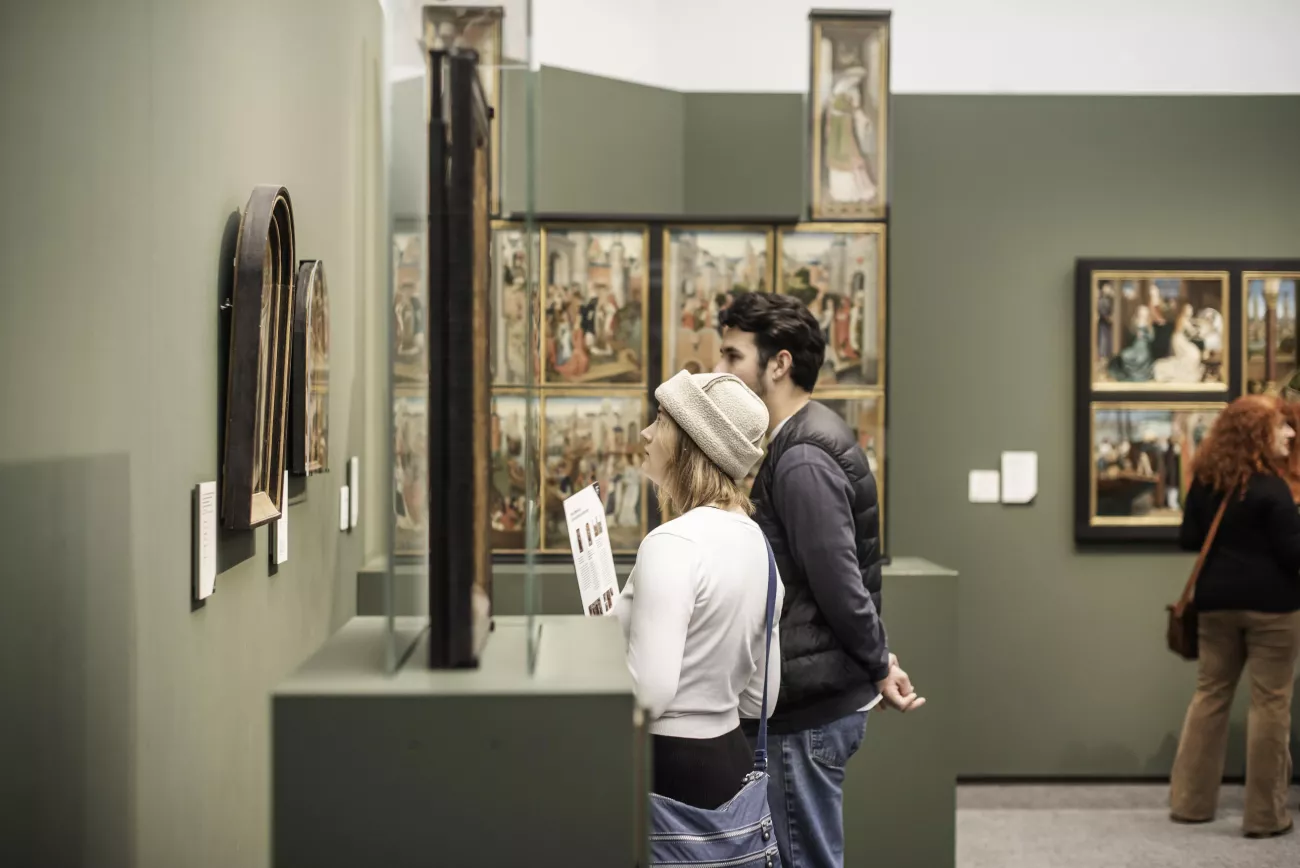 Visitantes de un museo observan pinturas flamencas primitivas enmarcadas y una obra en panel alta dentro de una vitrina en una sala de exposiciones con paredes verdes