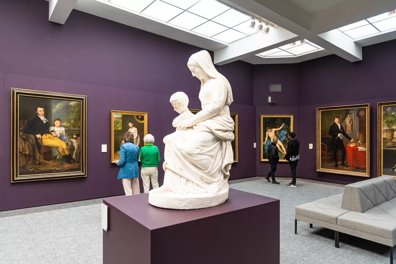 Interior de una galería de museo con paredes púrpuras que exhibe pinturas enmarcadas y una escultura de mármol blanco de una mujer sentada con un niño sobre un pedestal central, con visitantes observando el arte