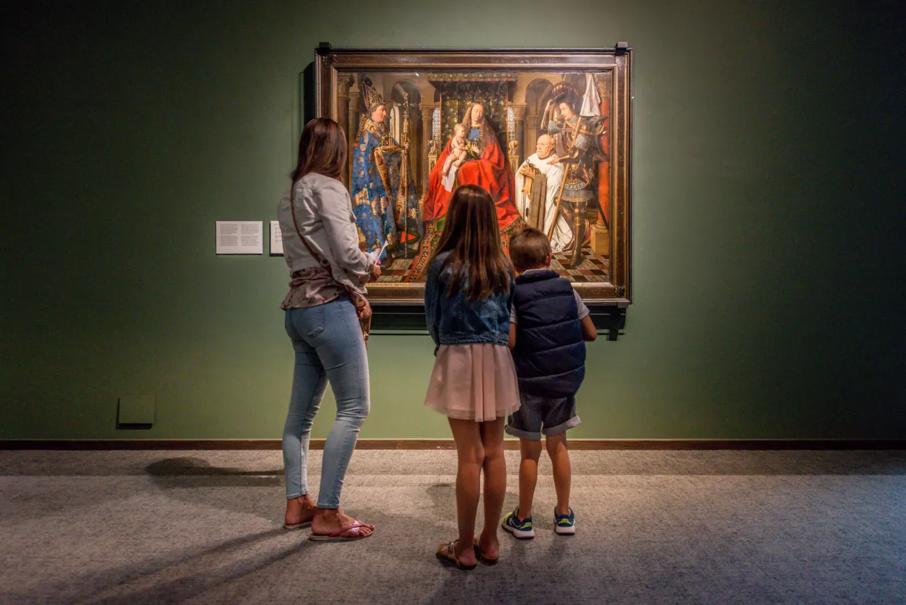 Tres visitantes están de pie frente a un gran cuadro religioso enmarcado que cuelga en una pared verde oscuro de la galería en el Groeningemuseum.