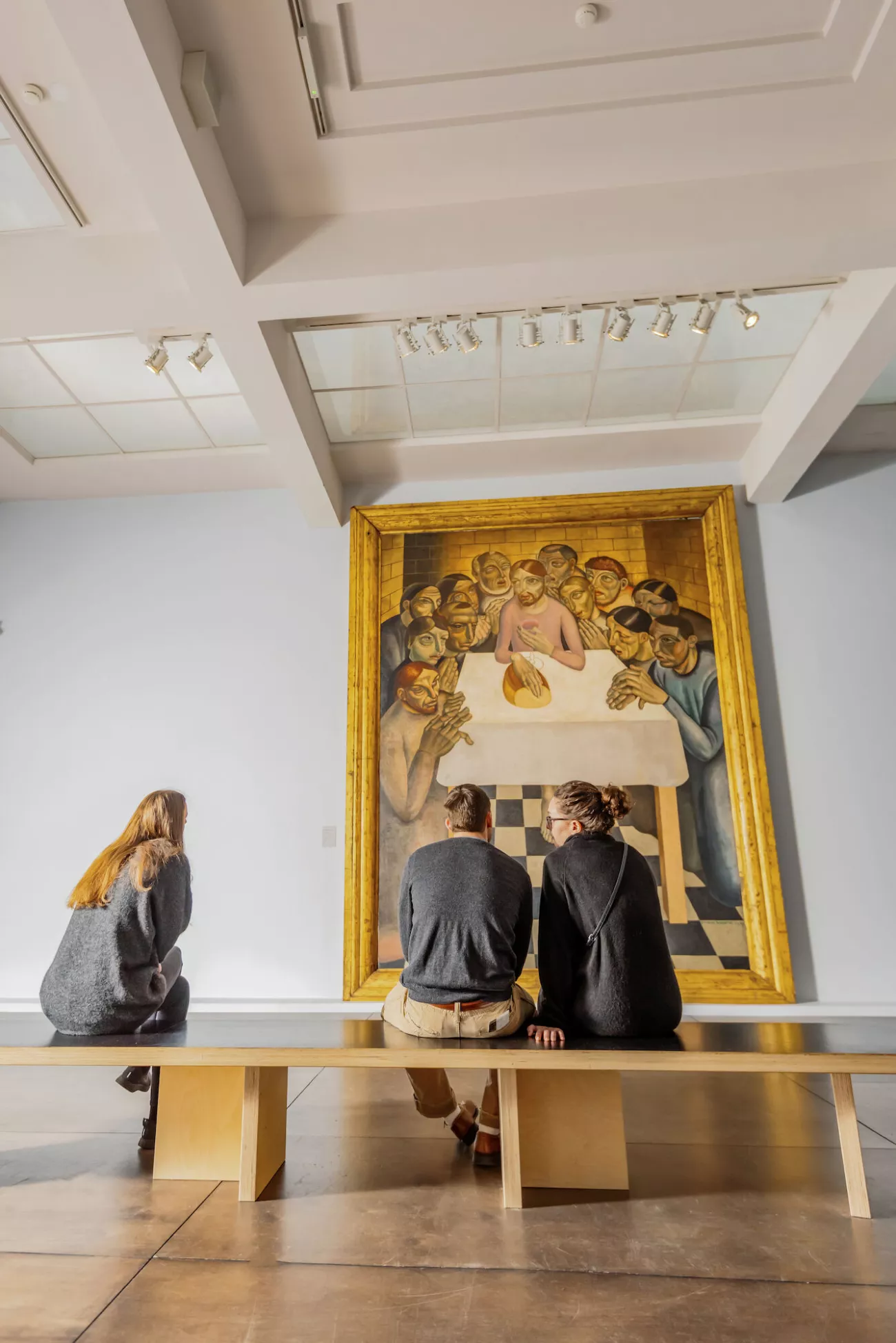 Tres visitantes sentados en un banco de madera observan un gran cuadro con marco dorado de figuras reunidas alrededor de una mesa en una galería de museo con iluminación de riel.