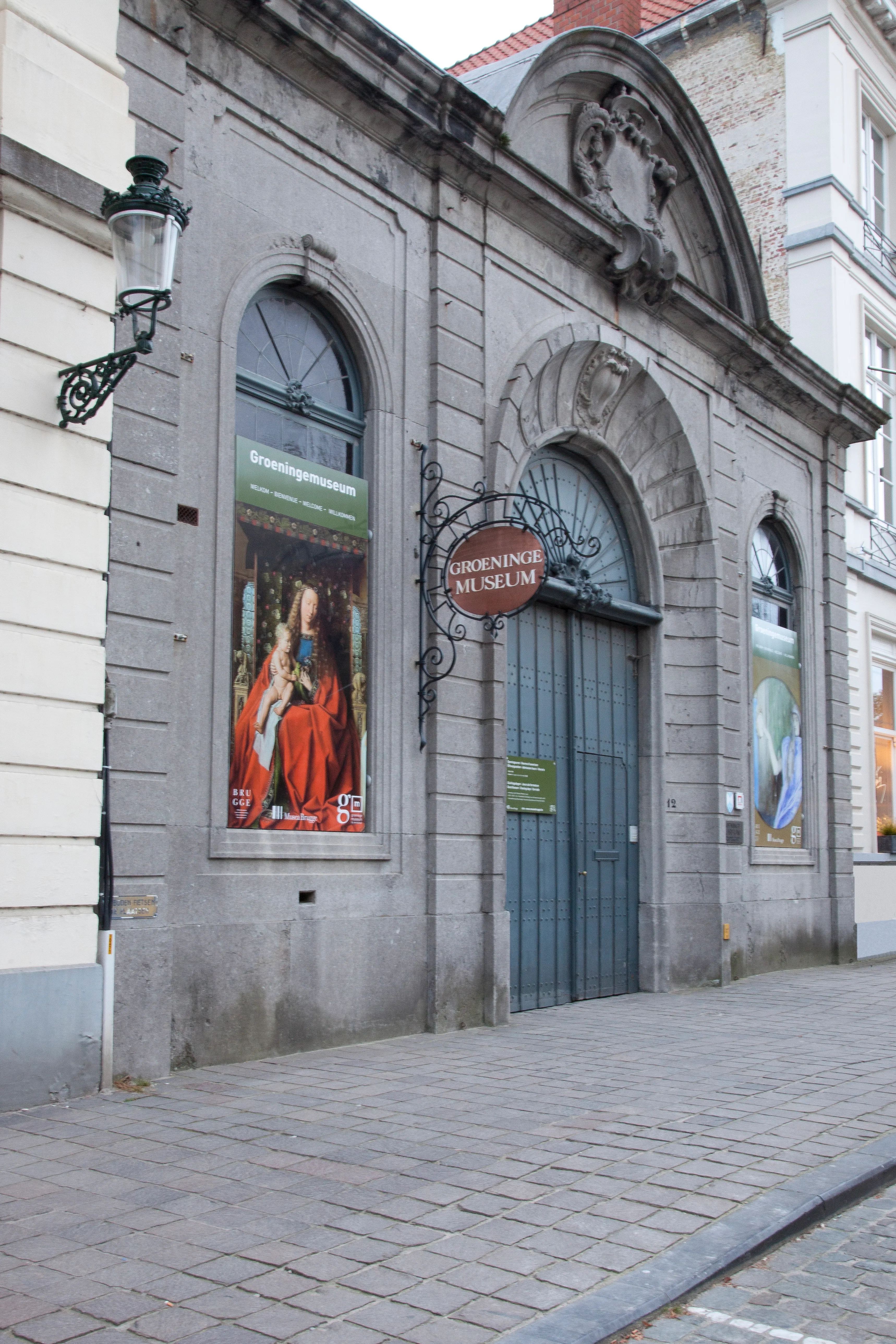 Kamienna fasada wejścia do Groeningemuseum z łukowatym wejściem, wiszącym owalnym szyldem z napisem 'Groeninge Museum' oraz plakatami wystawowymi w łukowatych oknach