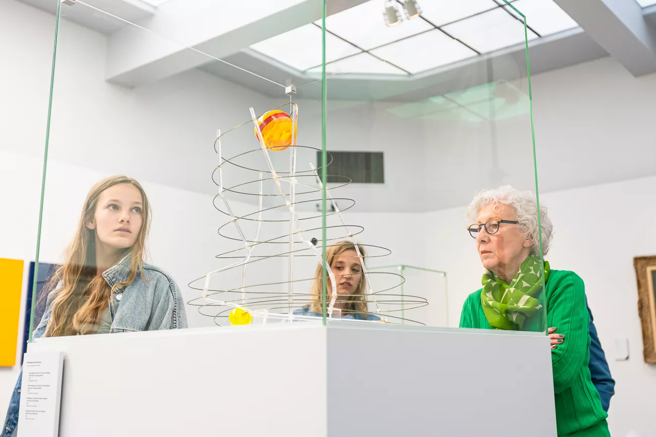 Tres visitantes observan una escultura cinética con cables metálicos en espiral y elementos coloridos dentro de una vitrina de cristal en una luminosa galería de museo con tragaluz.