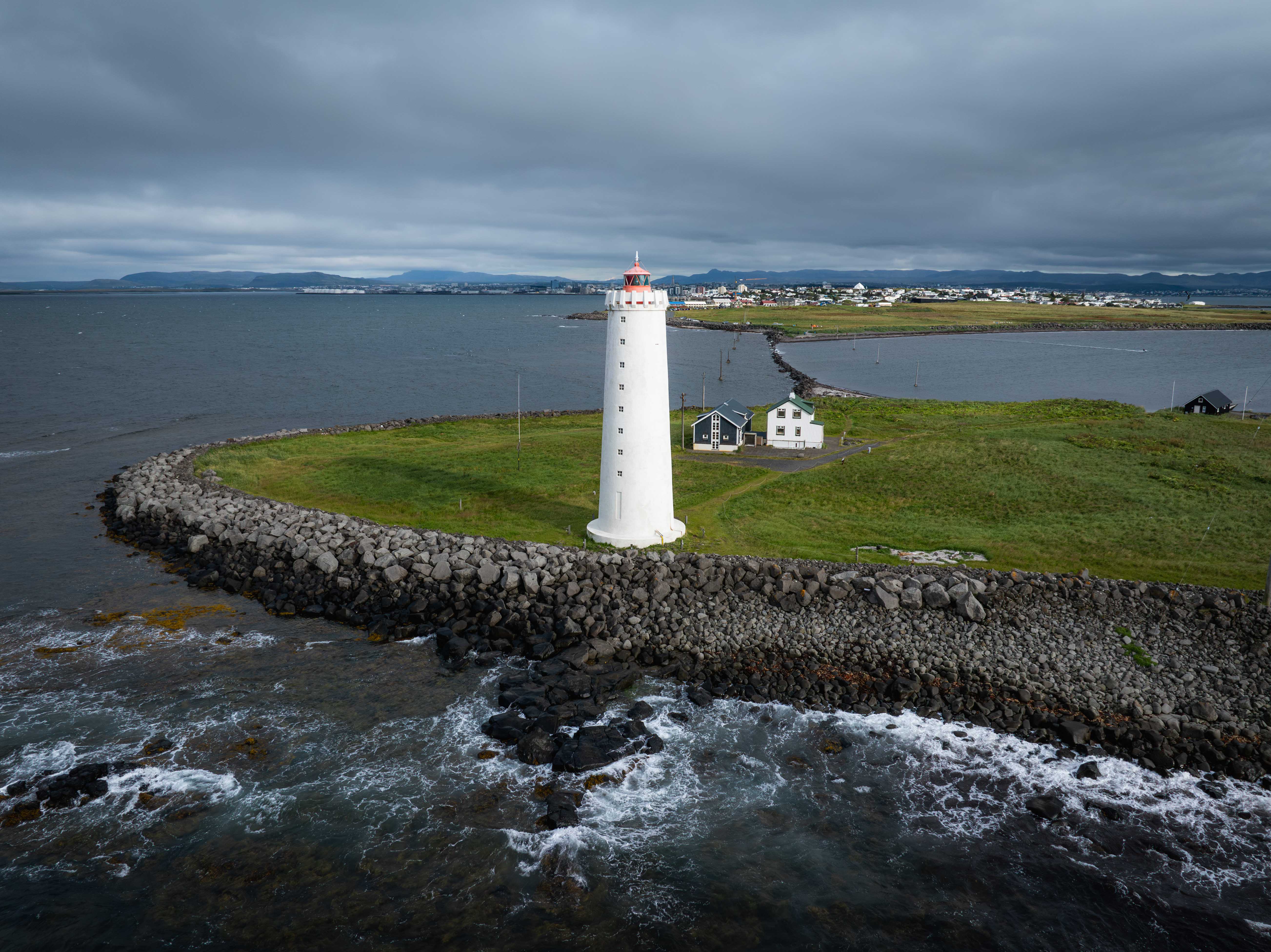 Faro cilíndrico blanco en una isla de marea cubierta de hierba, rodeado por un rompeolas de piedra, con olas del océano, cielo nublado y una ciudad distante al fondo.