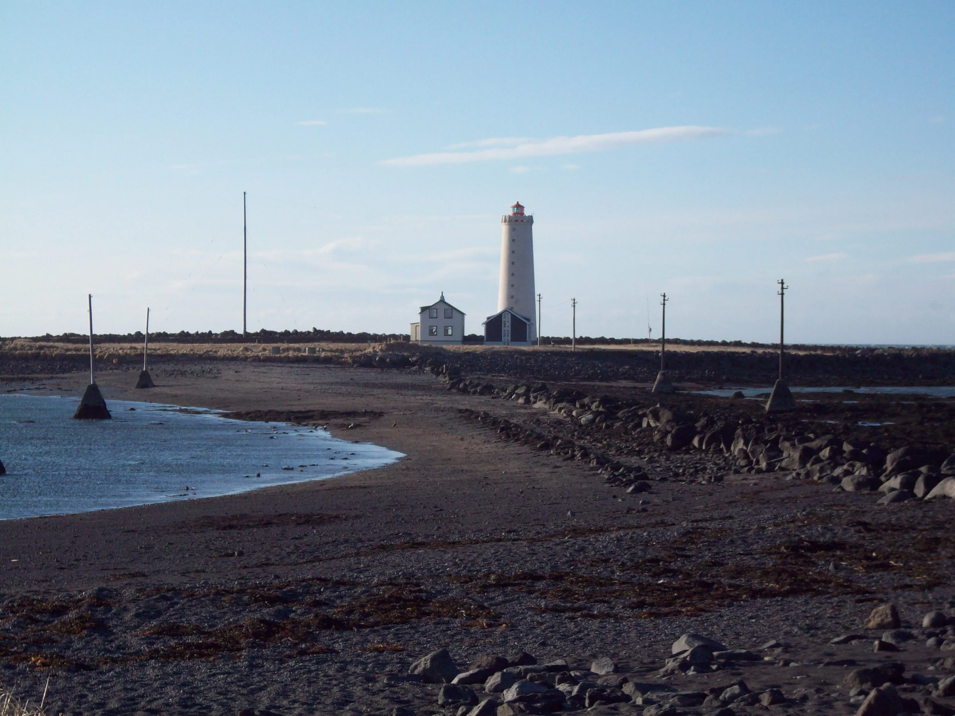 Un faro blanco y un pequeño edificio blanco se alzan sobre una isla rocosa, vistos desde una playa de arena con rocas dispersas y aguas tranquilas bajo un cielo azul.