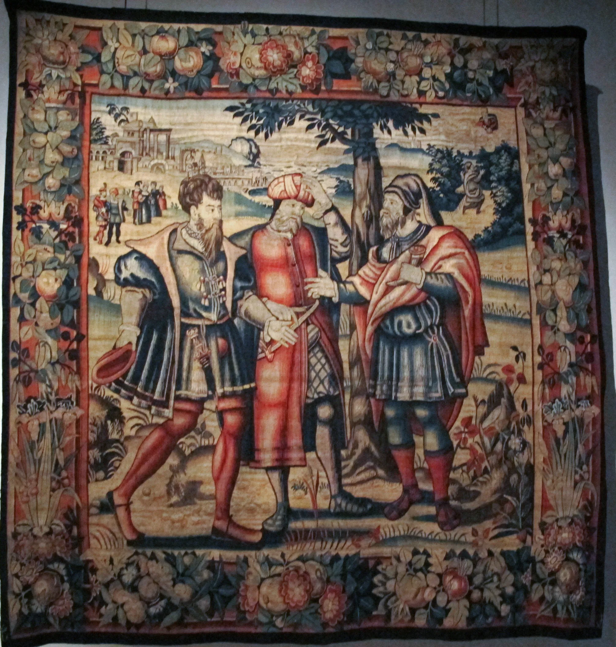 Un gran tapiz histórico que muestra a tres hombres con vestimentas renacentistas conversando al aire libre, rodeados por un denso borde floral.