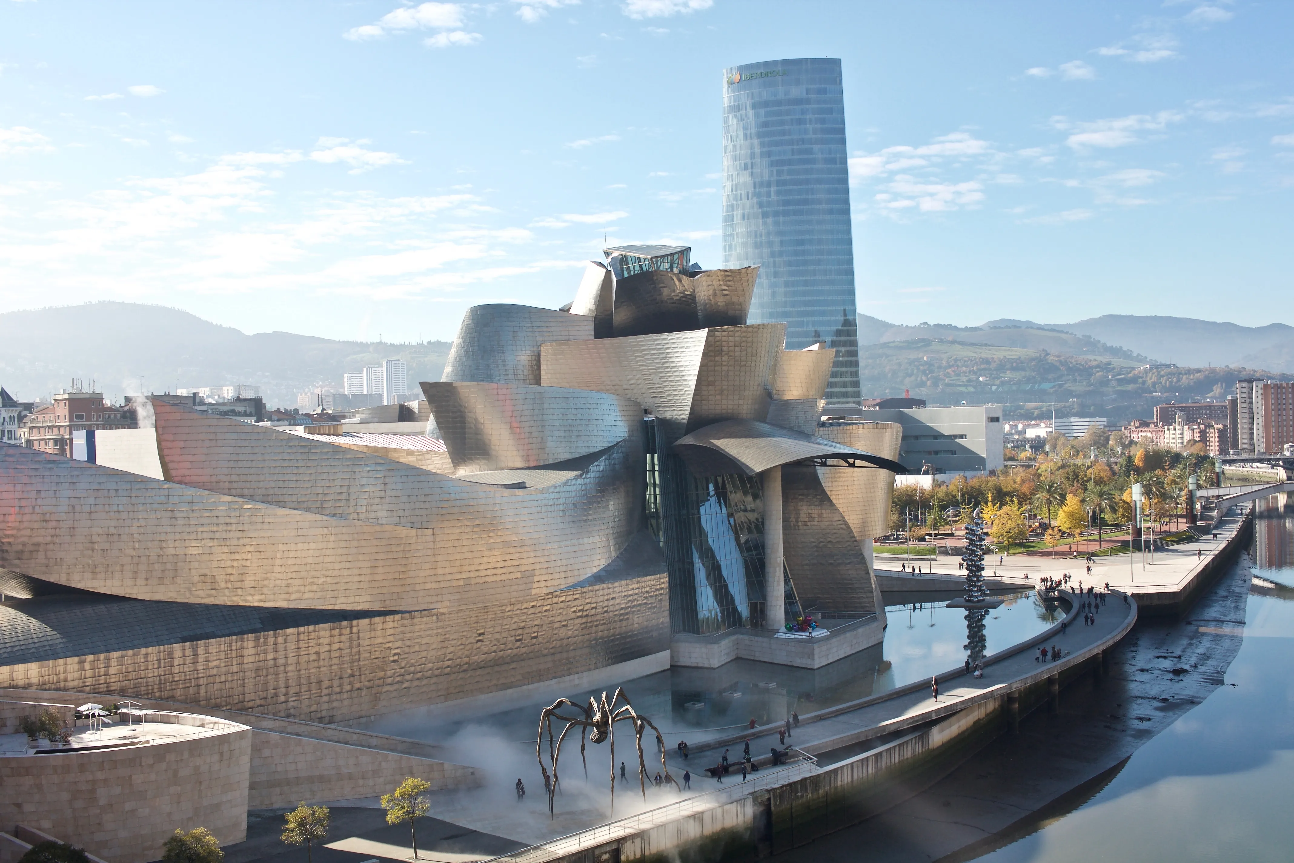 Widok z lotu ptaka na pokrytą tytanem, krzywoliniową architekturę Guggenheim Museum Bilbao wzdłuż rzeki Nervión, z rzeźbą pająka Maman i wieżowcem Iberdrola Tower w tle