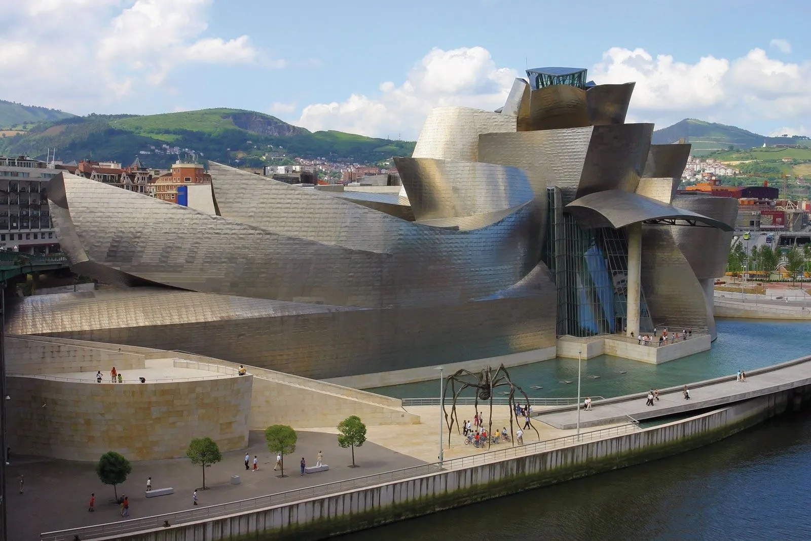Widok z lotu ptaka na Guggenheim Museum Bilbao, duży budynek pokryty tytanem o zakrzywionych kształtach przy rzece, ze wzgórzami w tle.