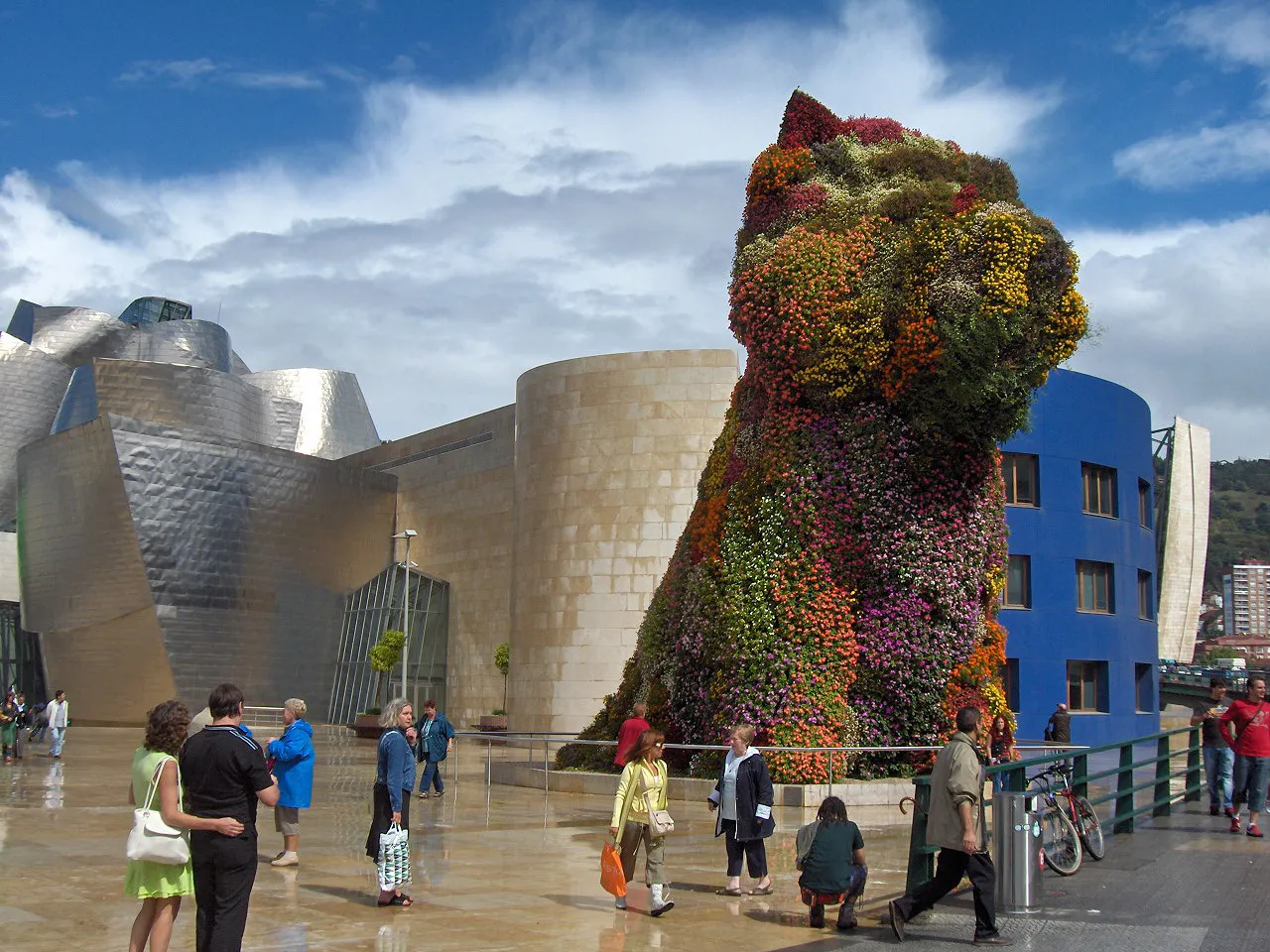 Rzeźba Puppy autorstwa Jeffa Koonsa przedstawiająca psa pokrytego kwiatami przed zakrzywionym budynkiem Guggenheim Museum Bilbao pokrytym tytanem, z odwiedzającymi spacerującymi po mokrym placu pod błękitnym niebem