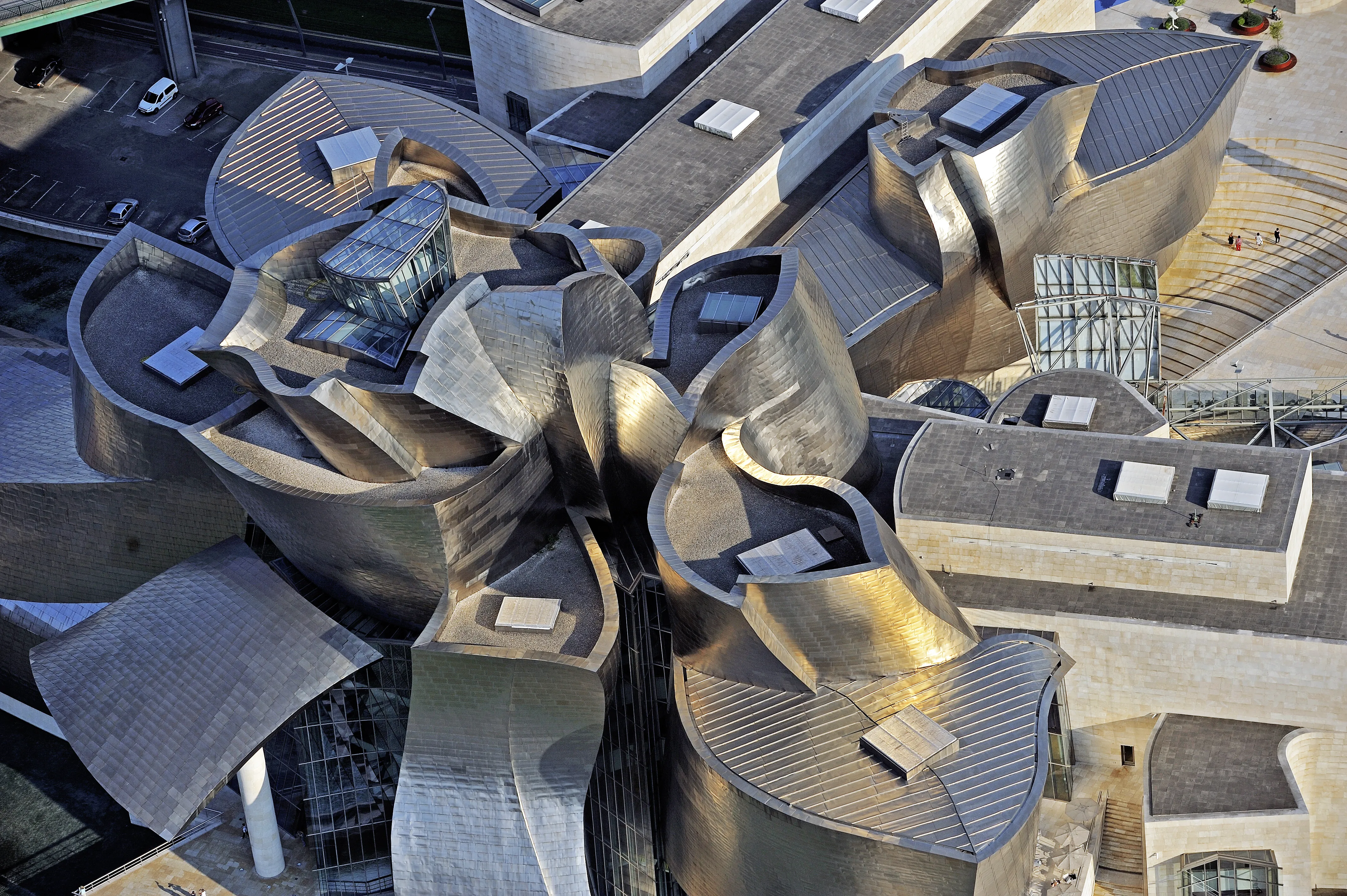 Widok z lotu ptaka na Guggenheim Museum Bilbao, ukazujący charakterystyczne, pokryte tytanem zakrzywione konstrukcje dachowe ułożone w kwiatowy wzór, z przeszklonymi świetlikami i metalicznymi powierzchniami.