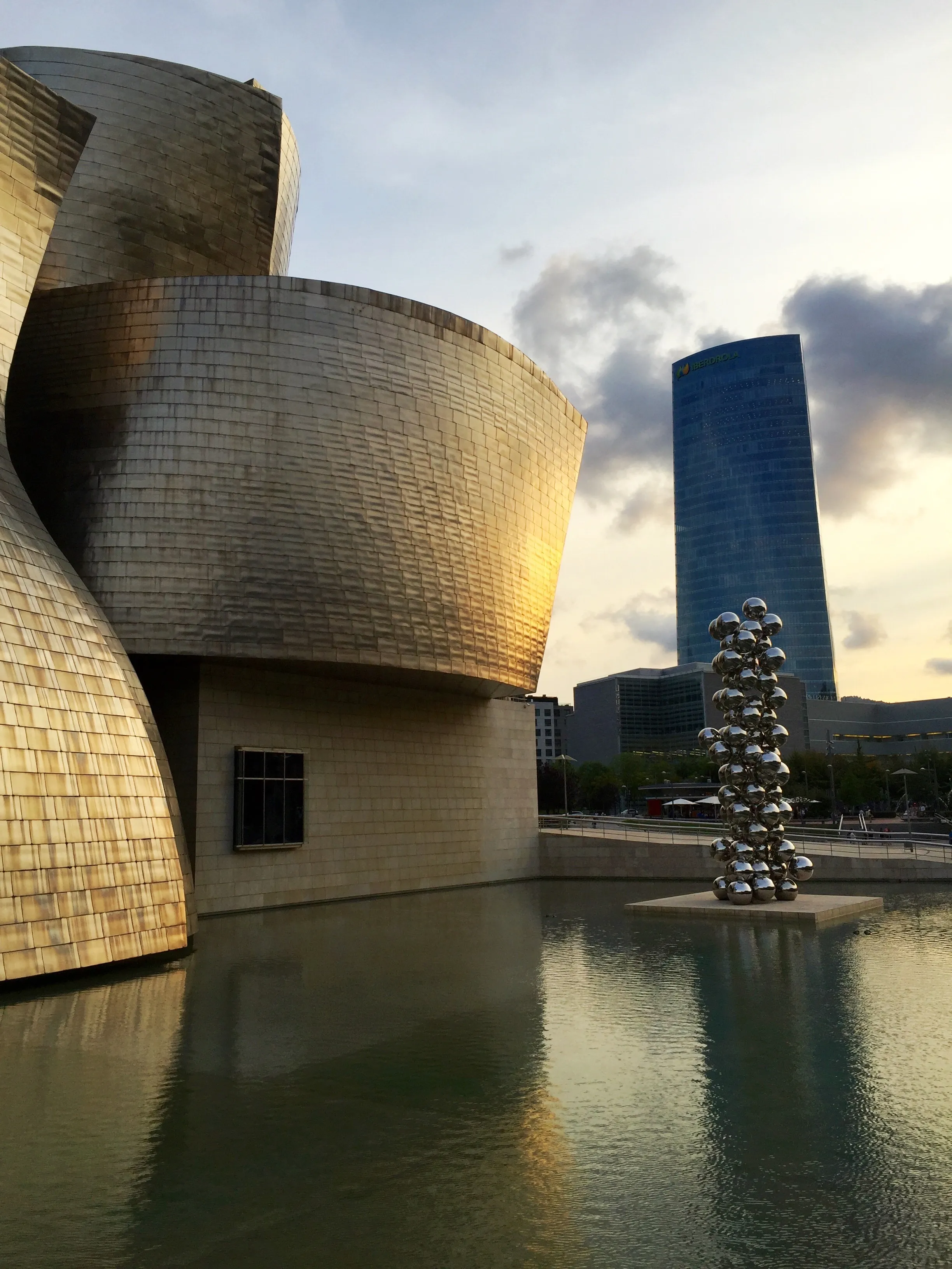 Zakrzywiona, pokryta tytanem fasada muzeum Guggenheim Museum Bilbao odbijająca się w tafli wody, ze stojącą w tle rzeźbą ze sterty metalowych kul i wysoką szklaną wieżą pod zachmurzonym niebem.