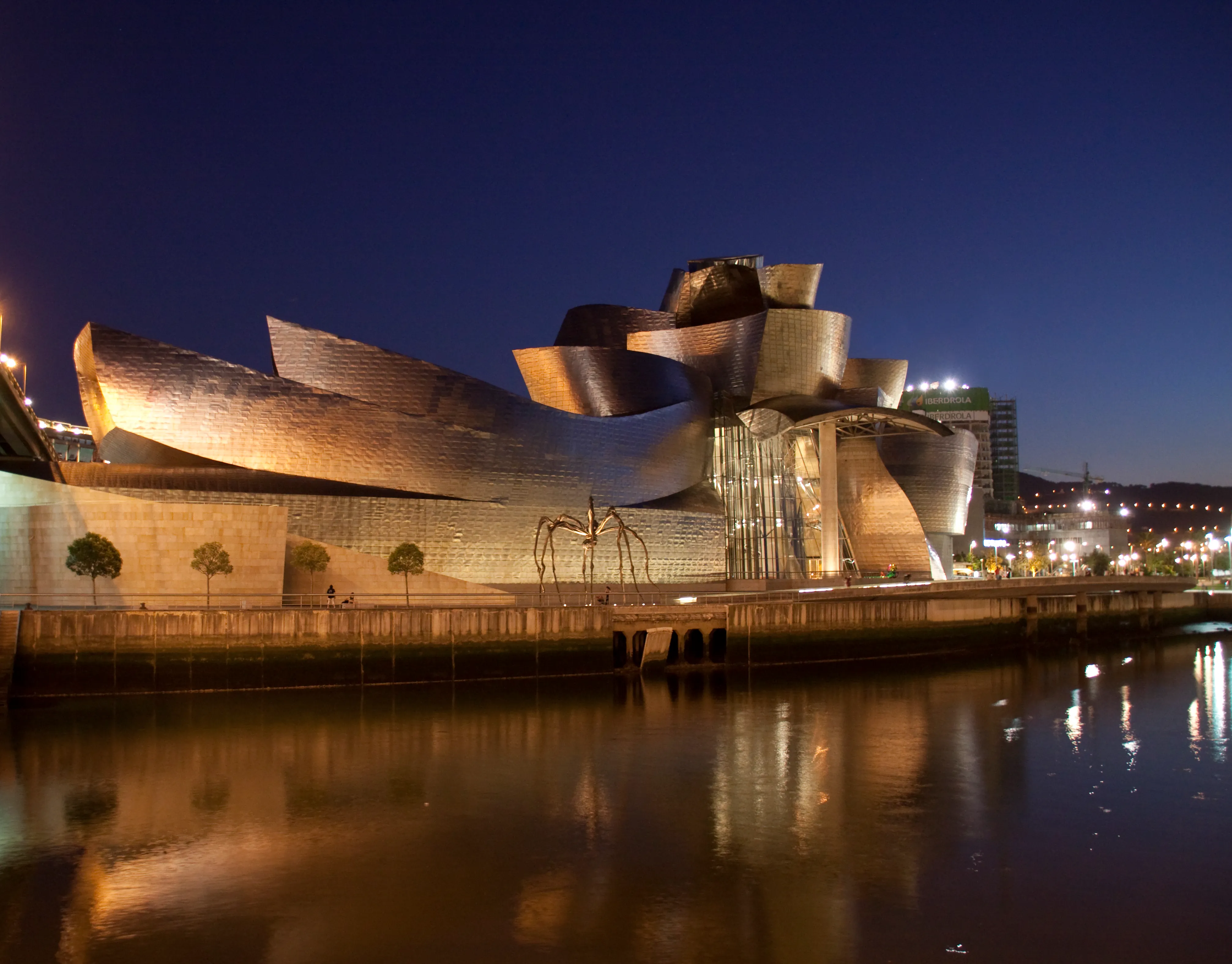 Guggenheim Museum Bilbao oświetlone nocą, z krzywiznami pokrytymi tytanem odbijającymi się w rzece Nervión