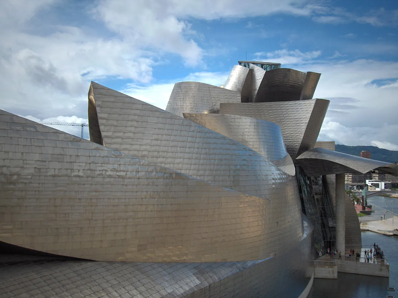 Zakrzywione, pokryte tytanem panele fasady Guggenheim Museum Bilbao na tle błękitnego nieba z rozproszonymi chmurami, z widocznymi w pobliżu wejścia zwiedzającymi i rzeką płynącą obok.