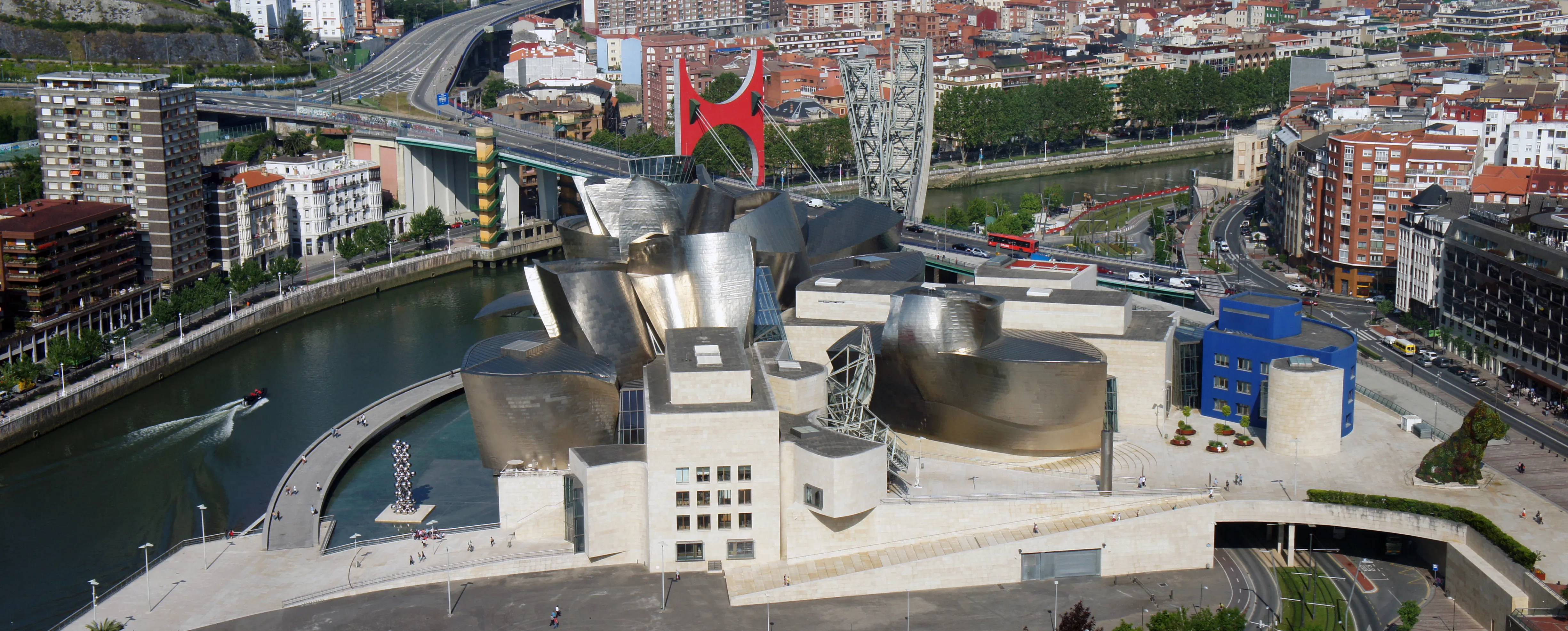 Panoramiczny widok z lotu ptaka na Muzeum Guggenheima w Bilbao z jego zakrzywioną, pokrytą tytanem architekturą wzdłuż rzeki Nervión, dużą czerwoną rzeźbą i otaczającym krajobrazem miejskim.