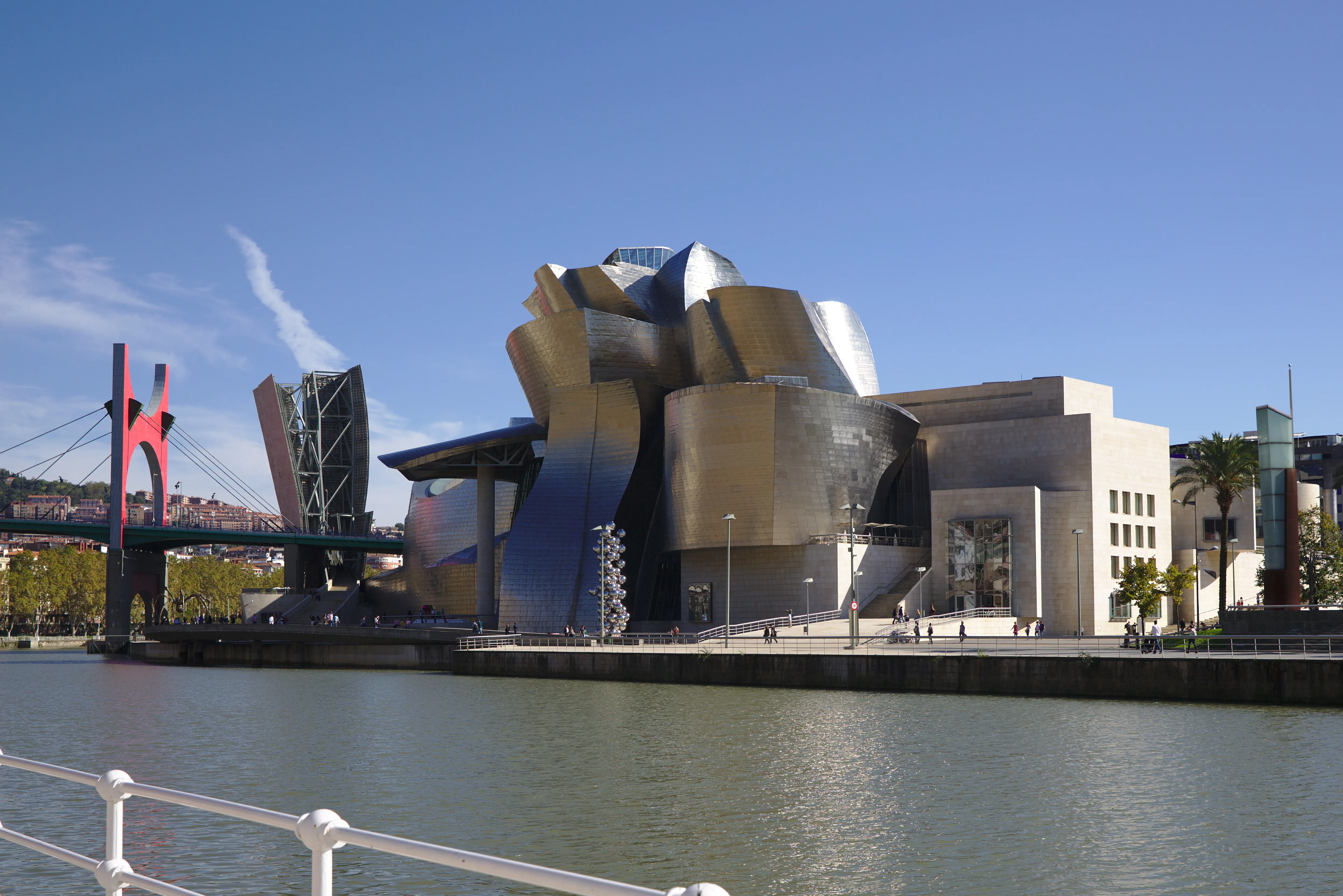Pokryty tytanem, krzywoliniowy budynek Muzeum Guggenheima w Bilbao widziany z drugiego brzegu rzeki Nervión, z czerwoną konstrukcją mostu widoczną po lewej stronie pod bezchmurnym, błękitnym niebem.