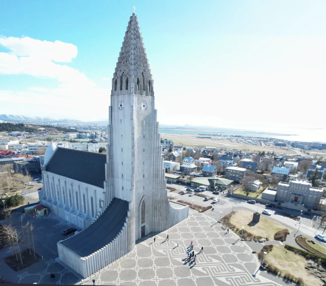 Luchtfoto van de Hallgrímskirkja-kerk met zijn hoge, op basalt geïnspireerde toren, het stenen plein met patronen en het stadsgezicht van Reykjavík met de kustlijn op de achtergrond.