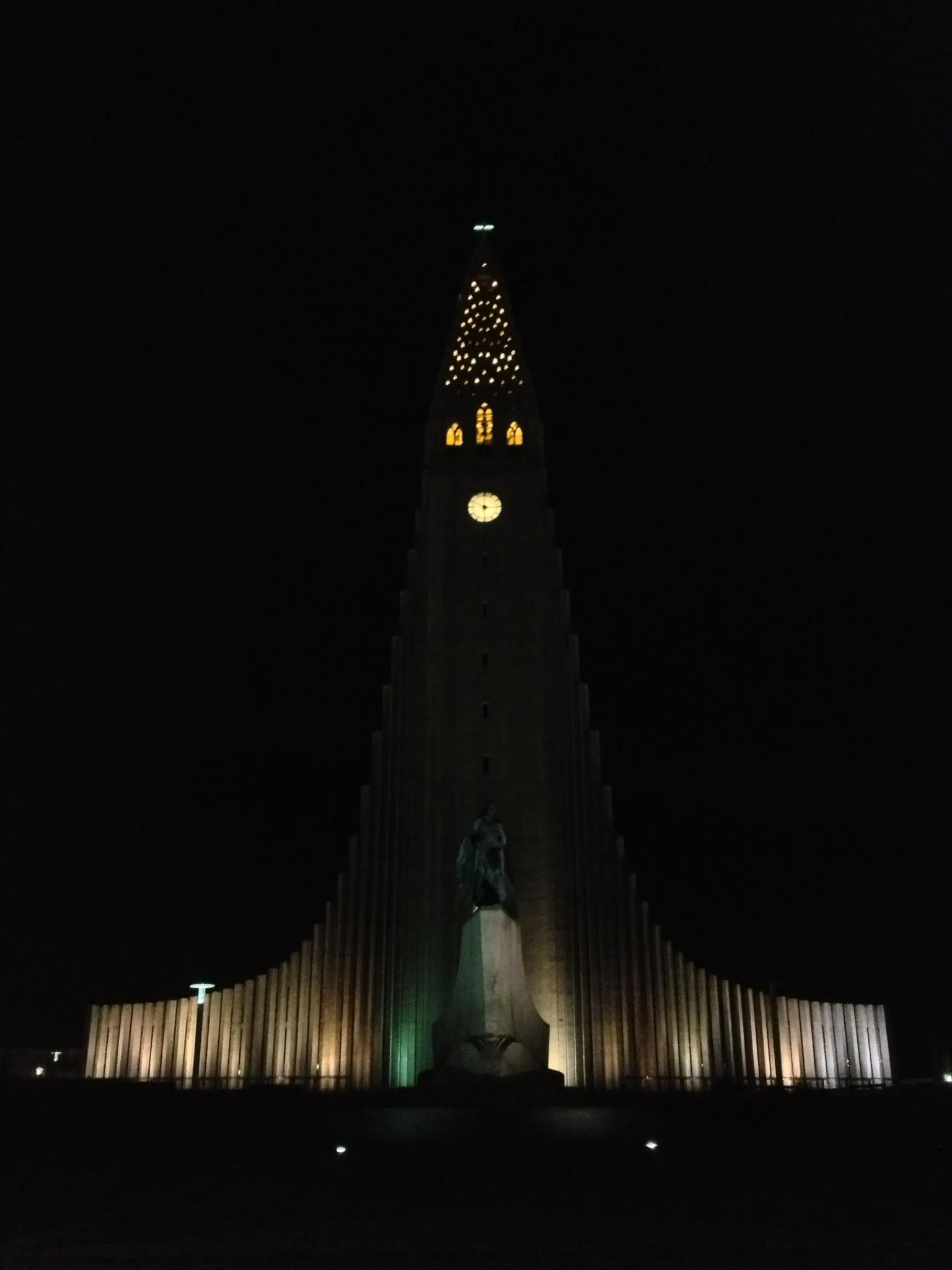 De kerktoren van de Hallgrímskirkja 's nachts verlicht, met zijn hoge spits, wijzerplaat en een standbeeld aan de voet.