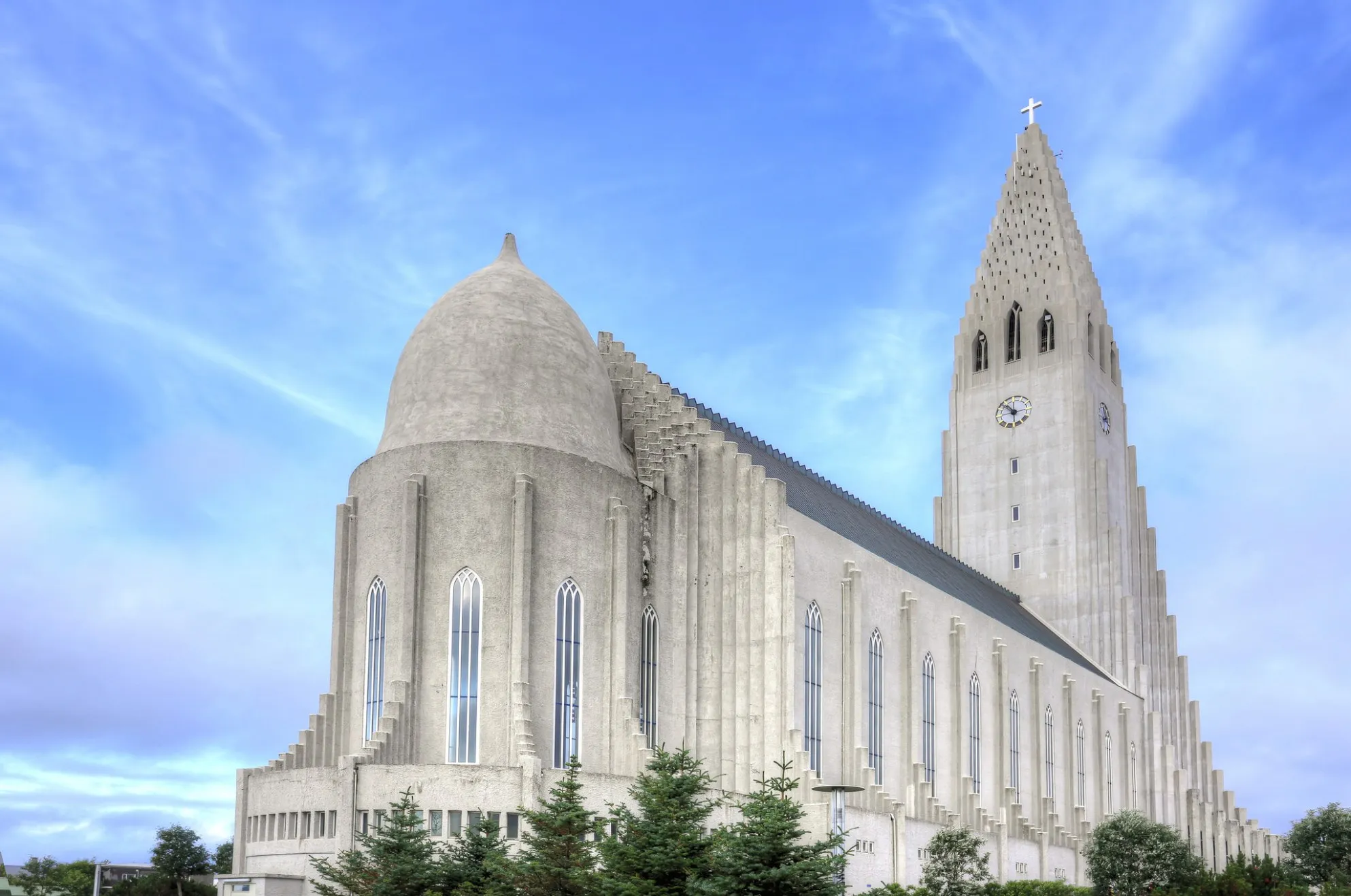 Zewnętrzna część kościoła Hallgrímskirkja z wysoką, schodkową wieżą z tarczą zegara i krzyżem, łukowymi oknami wzdłuż boku oraz błękitnym niebem w tle