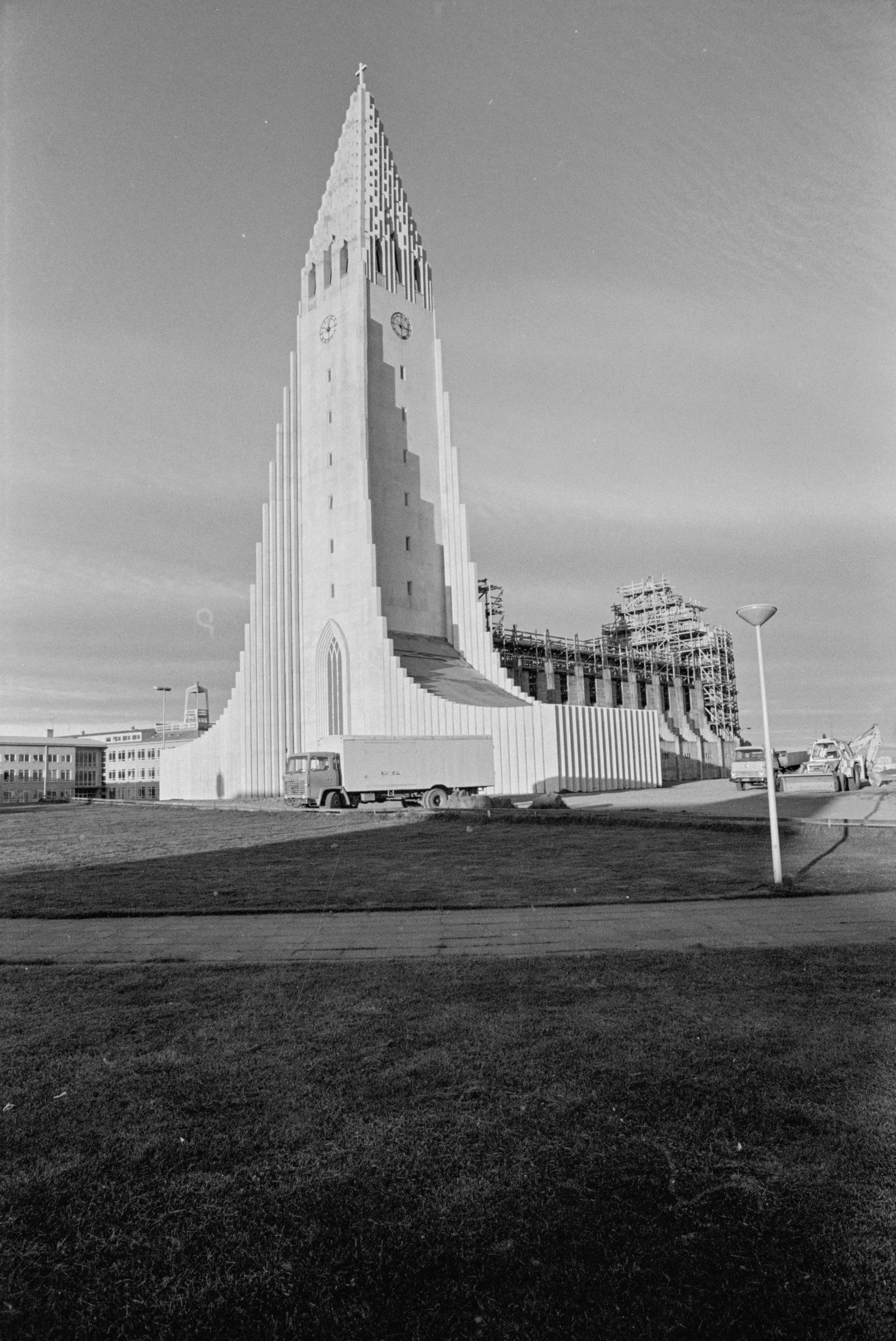 Zwart-witfoto van de toren van de Hallgrímskirkja in aanbouw, met steigers aan de rechterkant, een vrachtwagen geparkeerd aan de voet en een met gras begroeide voorgrond met omliggende gebouwen in de verte.