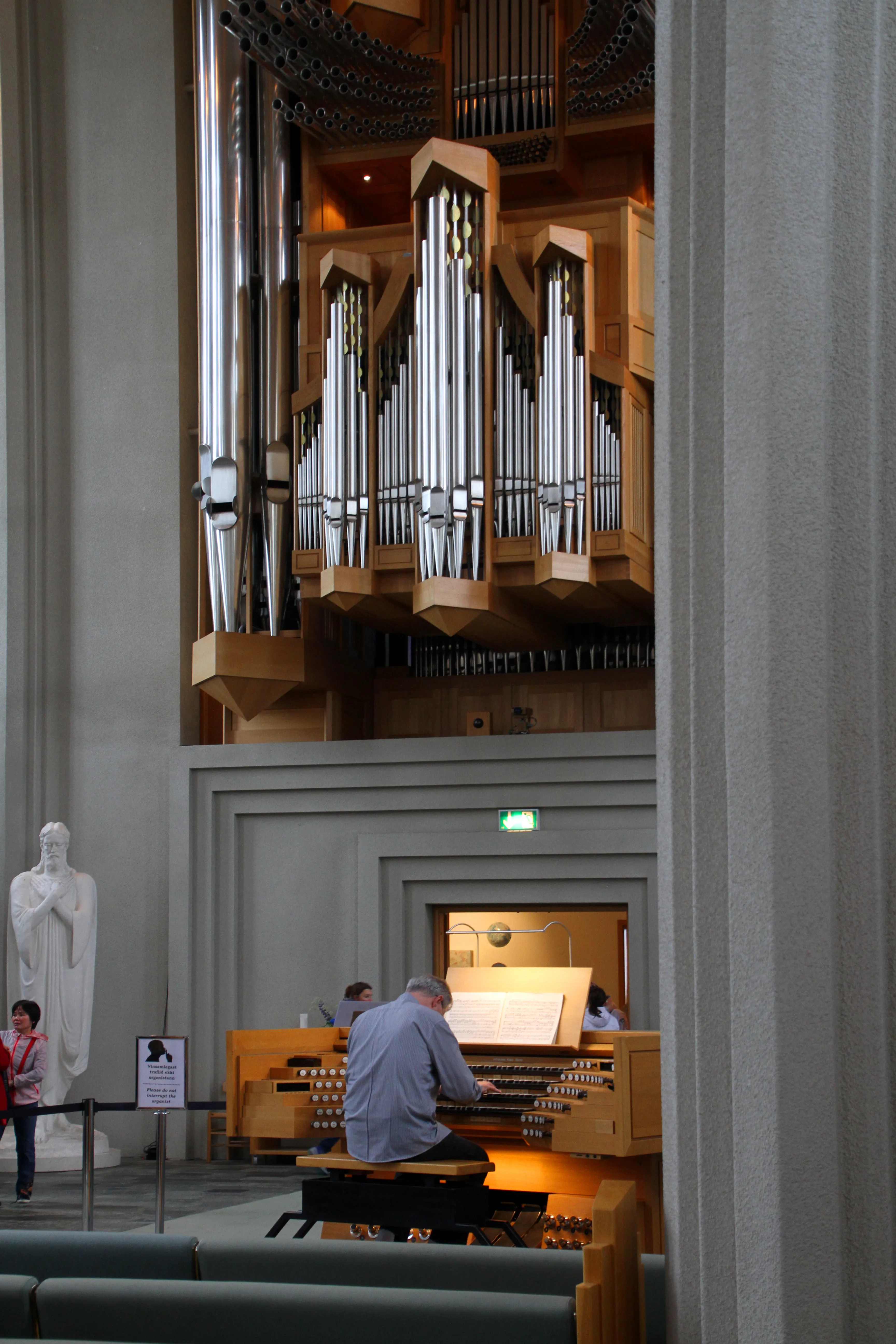 Groot pijporgel met houten omkasting en zilveren pijpen in de Hallgrímskirkja, met een organist aan de speeltafel en een wit standbeeld links zichtbaar.