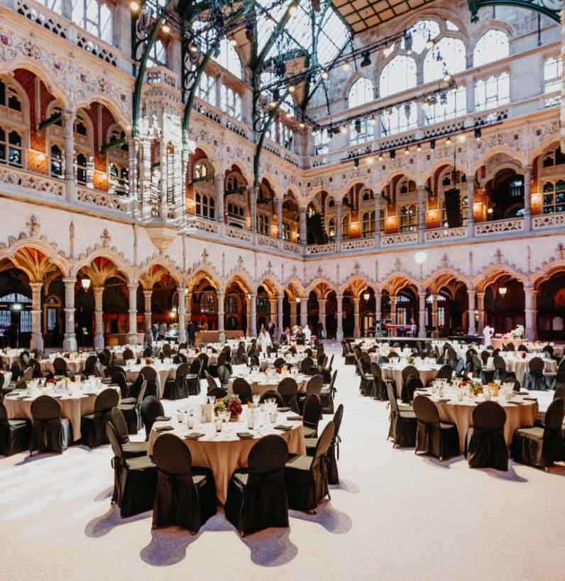Gran salón neogótico con galerías arqueadas ornamentadas y techo de cristal, dispuesto con mesas redondas de banquete y sillas negras.