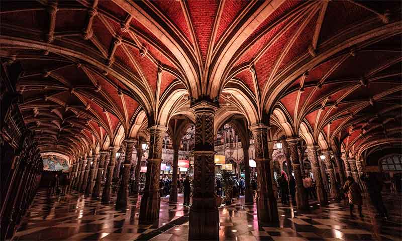 Techo abovedado neogótico de ladrillo rojo con nervaduras de piedra, sostenido por columnas ornamentadas en la galería porticada de la Handelsbeurs Antwerpen