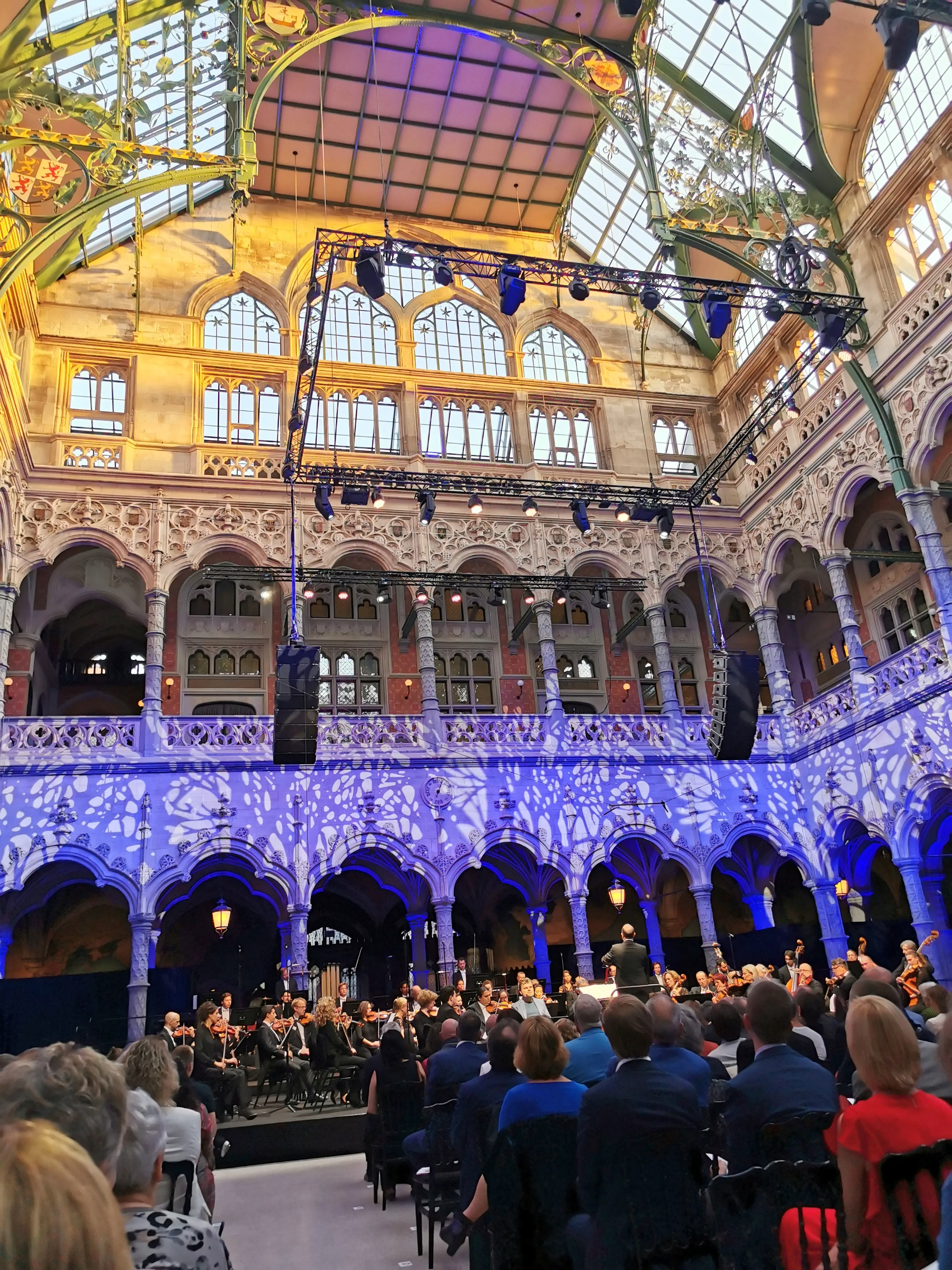 Gran salón interior neogótico con techo de cristal ornamentado, galerías arqueadas y proyecciones de luz azul en las paredes, con una orquesta actuando en el escenario y el público sentado en primer plano
